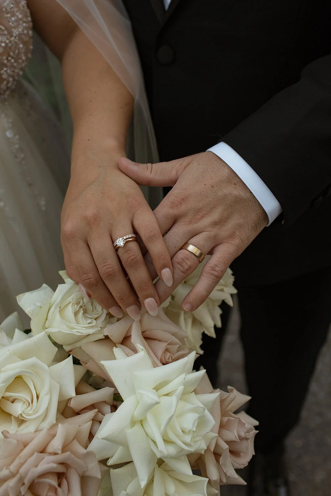 Close-up of a newlywed couple's hands showing wedding rings, with a bouquet of white and blush roses in the foreground.