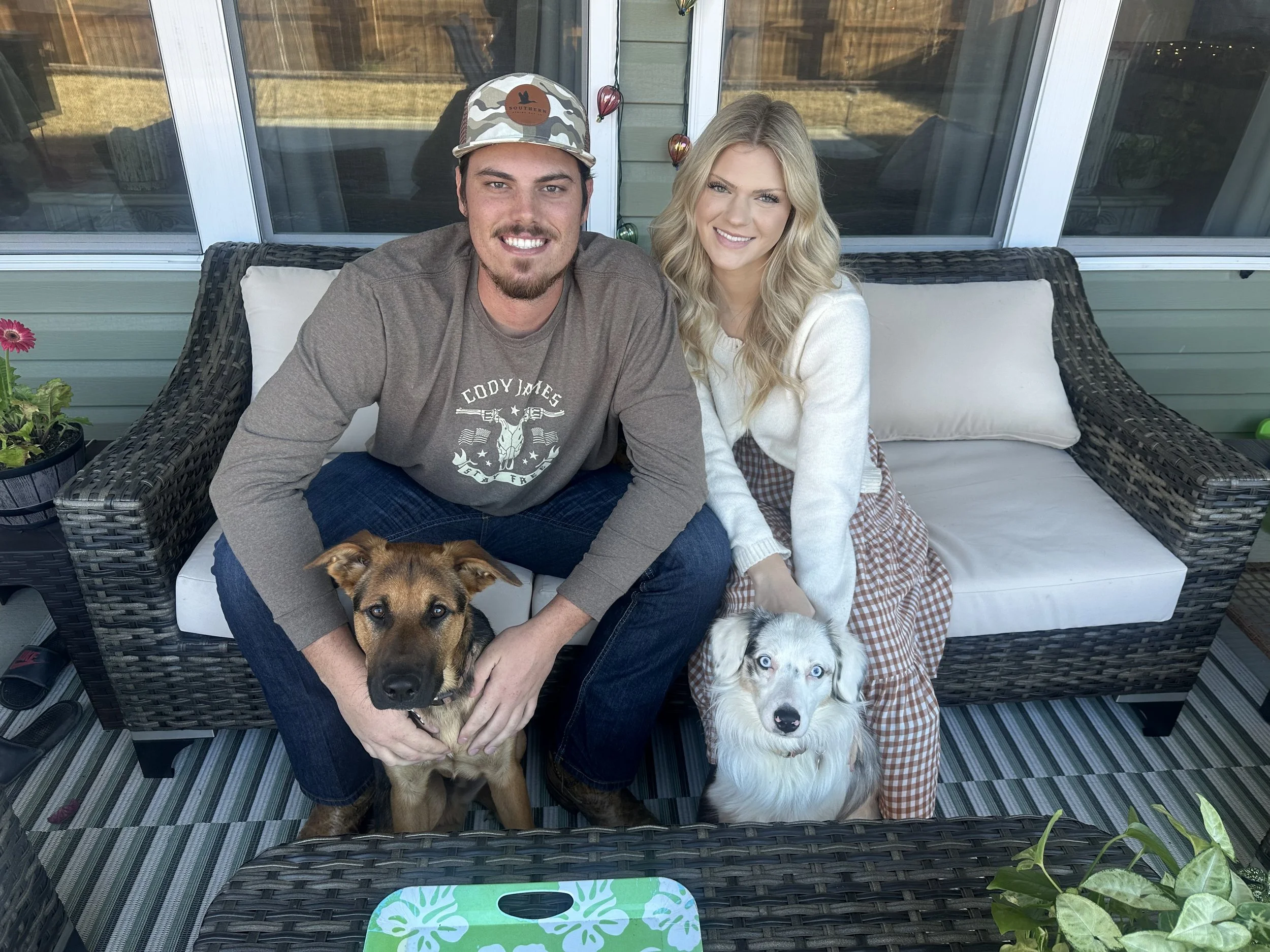 A smiling man with a beard and a backward cap sitting on a porch, holding a dog. Next to him, a smiling woman with blonde, wavy hair, in a white sweater and checkered skirt, sitting on a wicker outdoor sofa. Two dogs, one with brown and black fur and one with white and black fur, sit in front of them. The porch has a glass door, and a potted plant with pink flowers is visible on the left side. There are colorful string lights hanging behind.