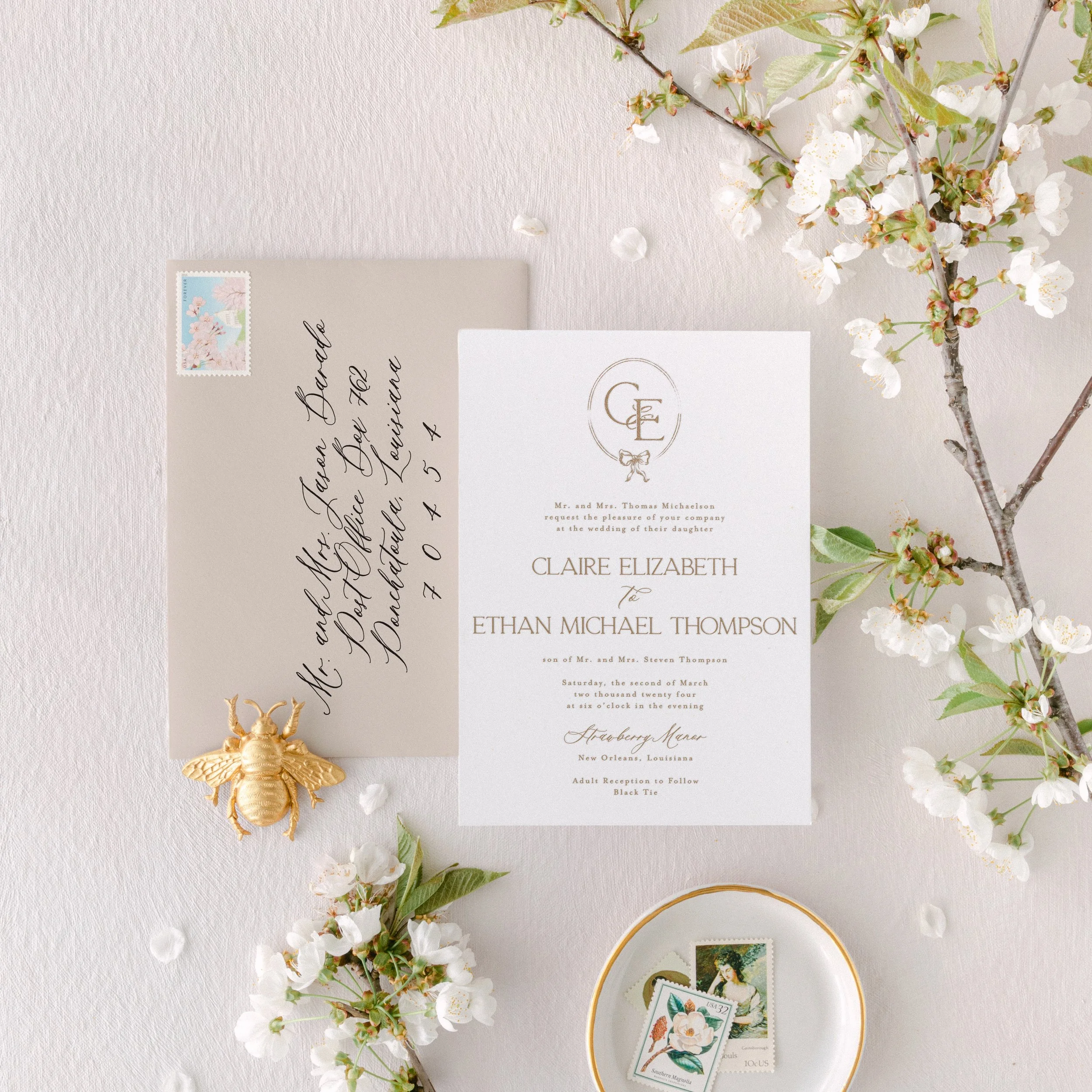 Wedding invitation set with elegant calligraphy, featuring an invitation card and a decorated envelope. Accompanied by cherry blossoms, a small golden bee ornament, and a dish with postage stamps. The arrangement is on a textured, light-colored surface.