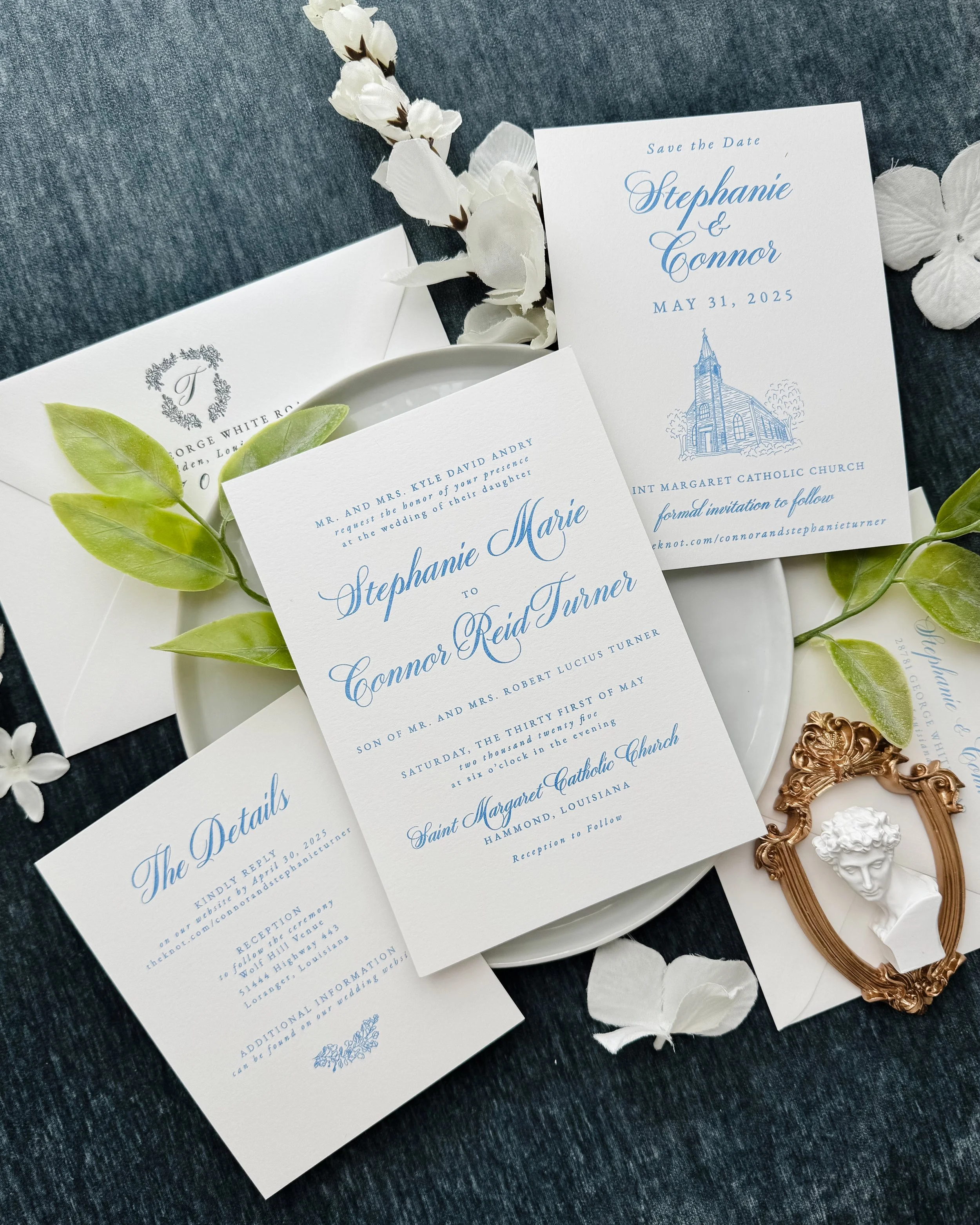 Wedding invitation suite with floral decorations, including an invitation, a save-the-date card, and an RSVP card, along with a small decorative bust in a gold frame and white petals scattered around, set on a dark fabric background.