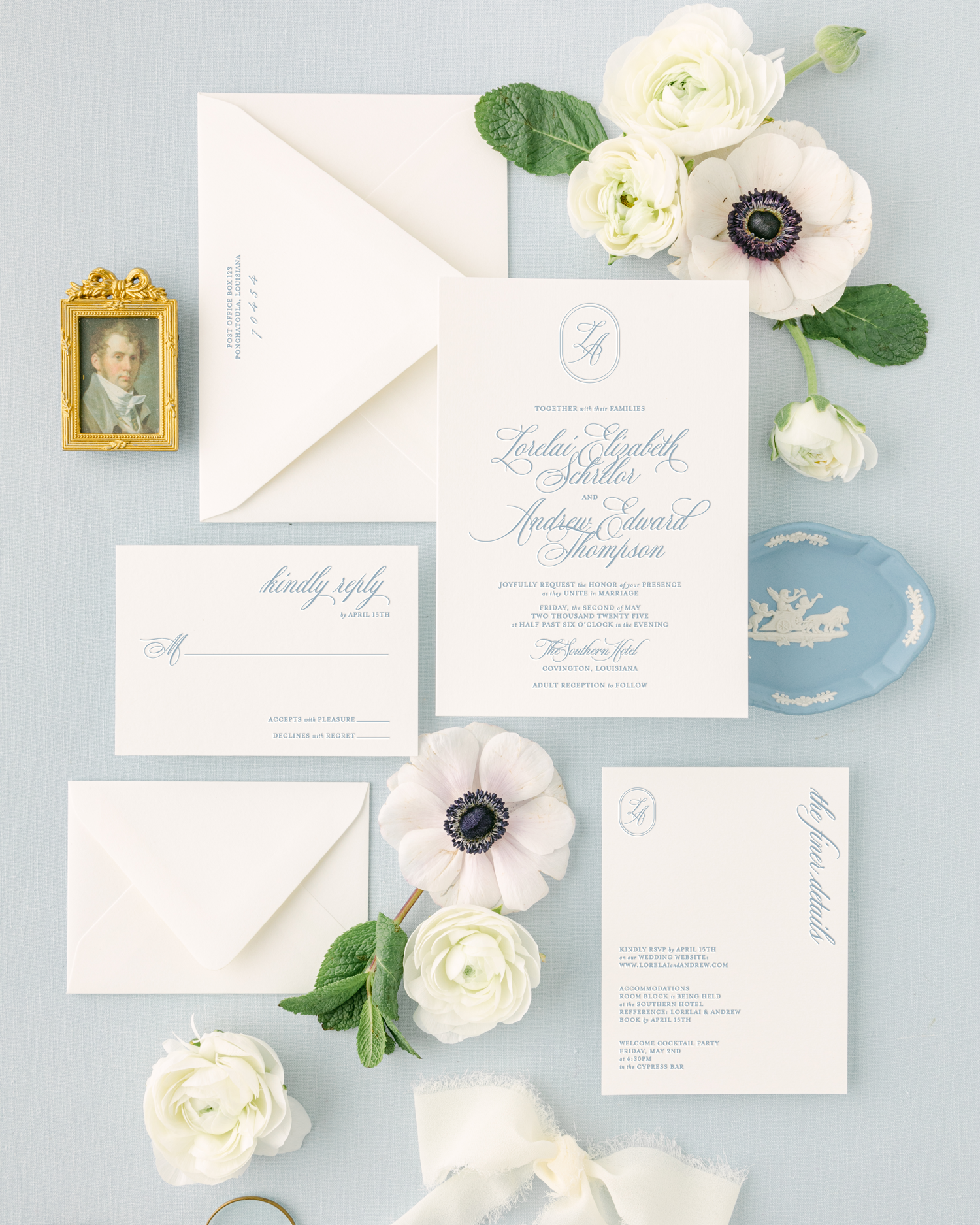 Wedding invitation suite with floral accents, including an invitation card, RSVP card, thank you card, white roses, an anemone flower, a small gold framed portrait, a blue porcelain dish, and white envelopes on a light blue background.