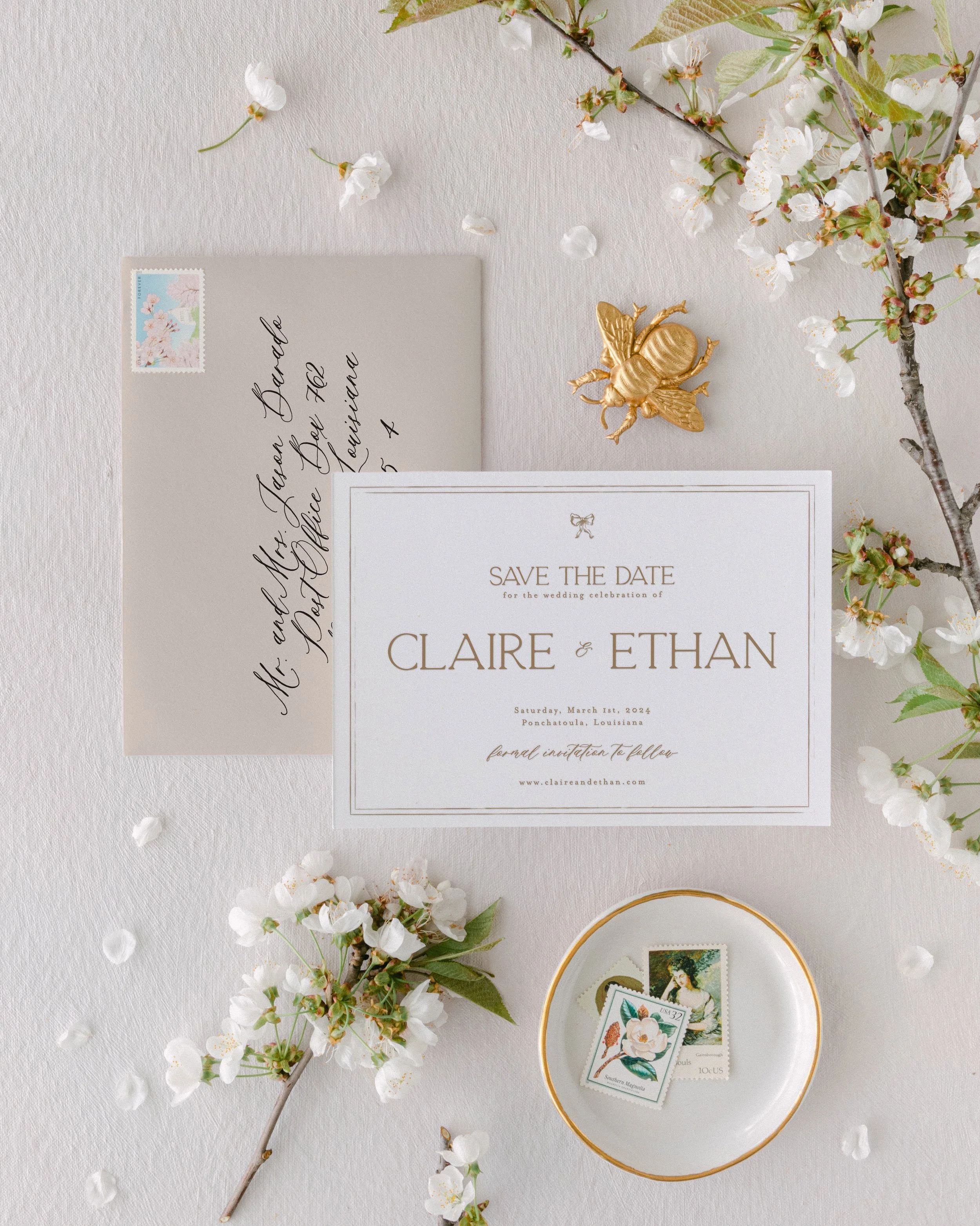 Save the date card for Claire and Ethan with envelope, postage stamps, flowering branches, and a gold insect decoration on a textured white surface.
