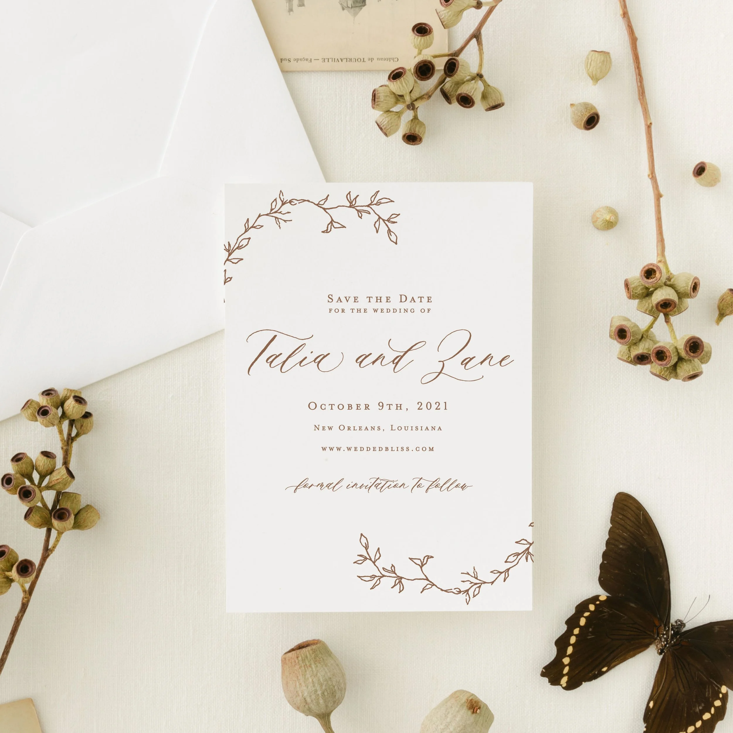 A wedding save the date card with floral illustrations, surrounded by dried seed pods, a white envelope, and a black butterfly, all on a light-colored fabric background.