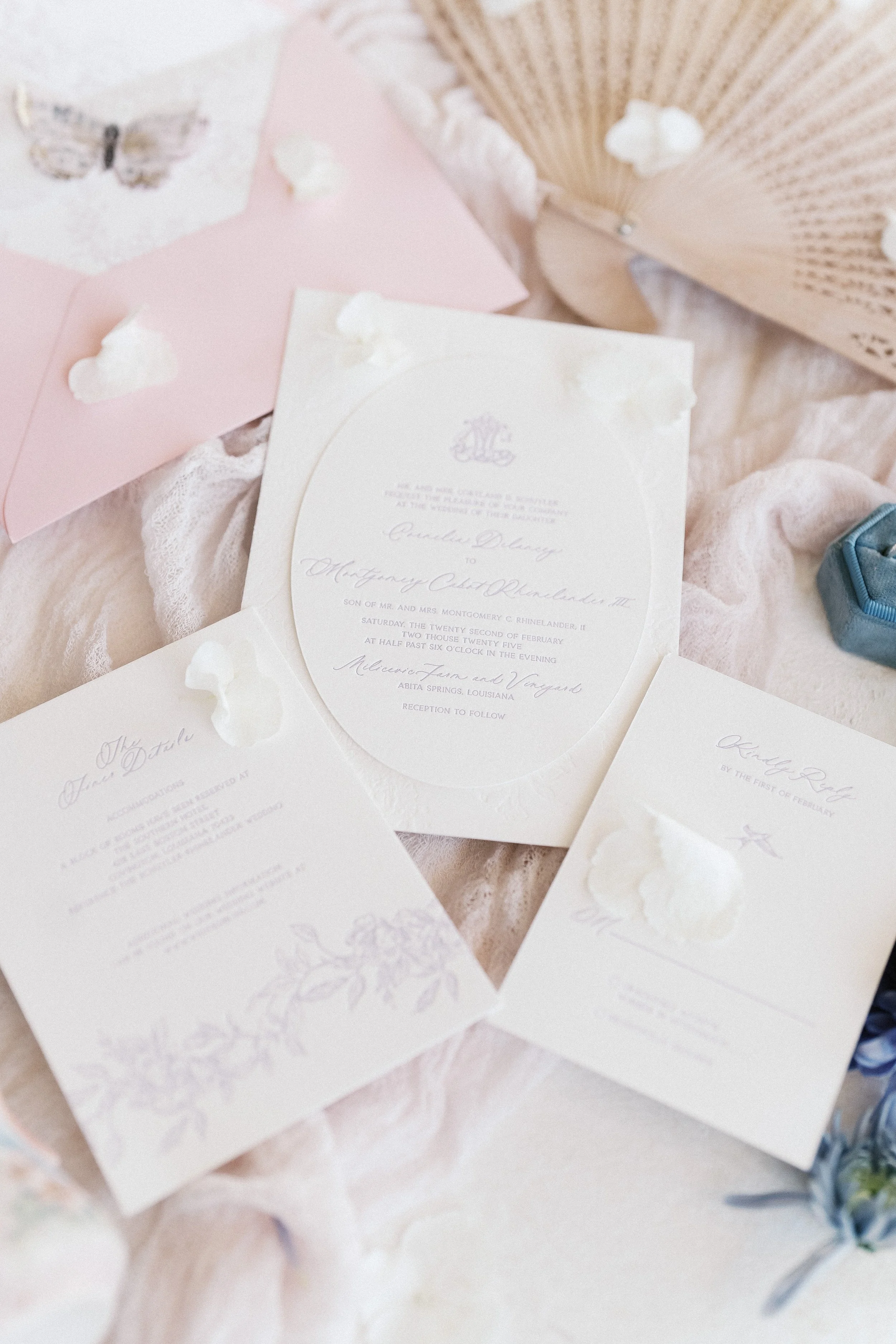 Elegant wedding invitations with floral designs, lace details, and script text, placed on a soft fabric with decorative elements like a pink envelope, a fan, and petals.