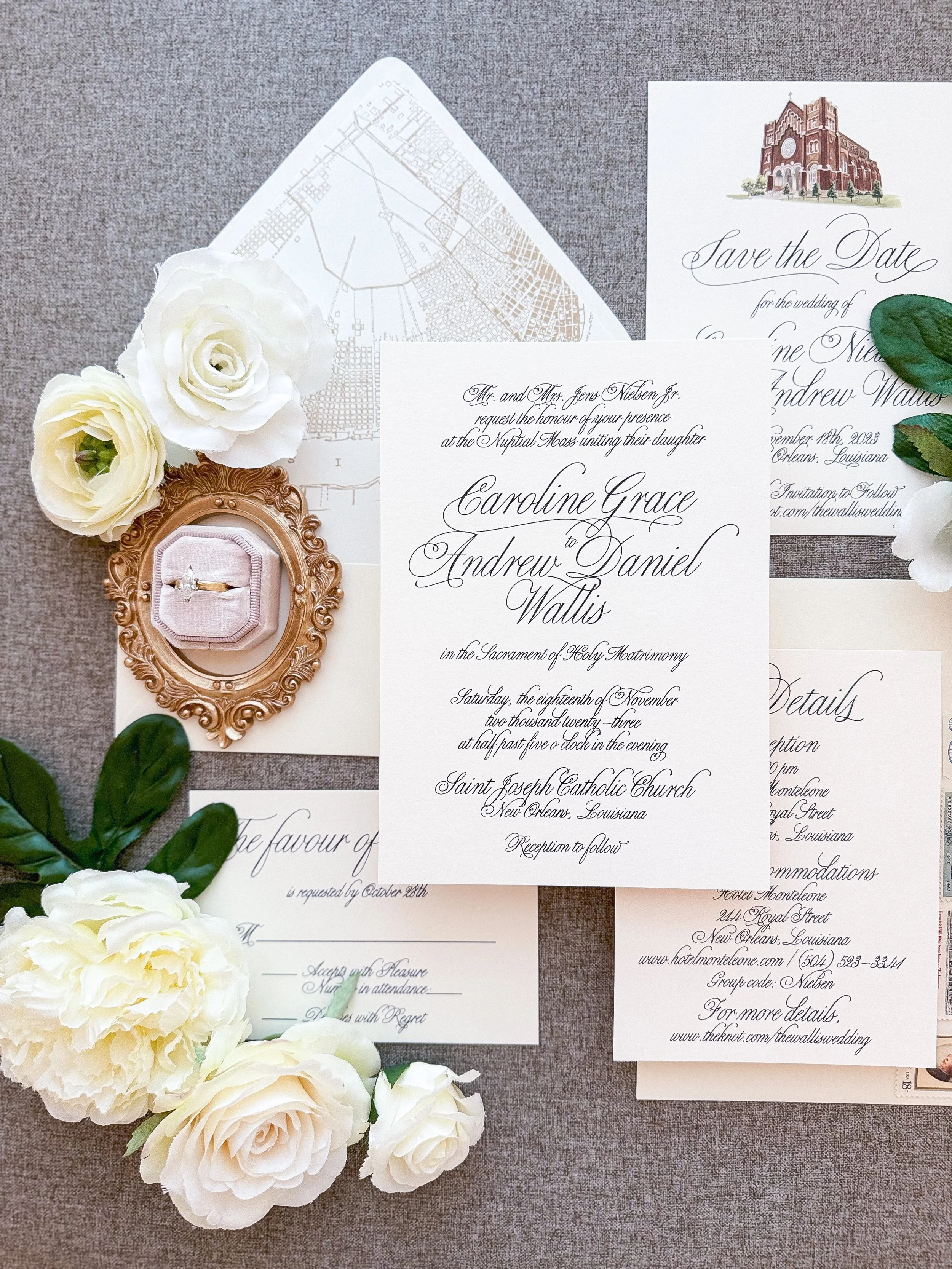 Flat lay image of wedding invitations with calligraphy text, surrounded by white roses and greenery. A gold-framed ring box with a ring is included, and the invitations show details about a wedding at Saint Joseph Catholic Church in New Orleans, Louisiana.