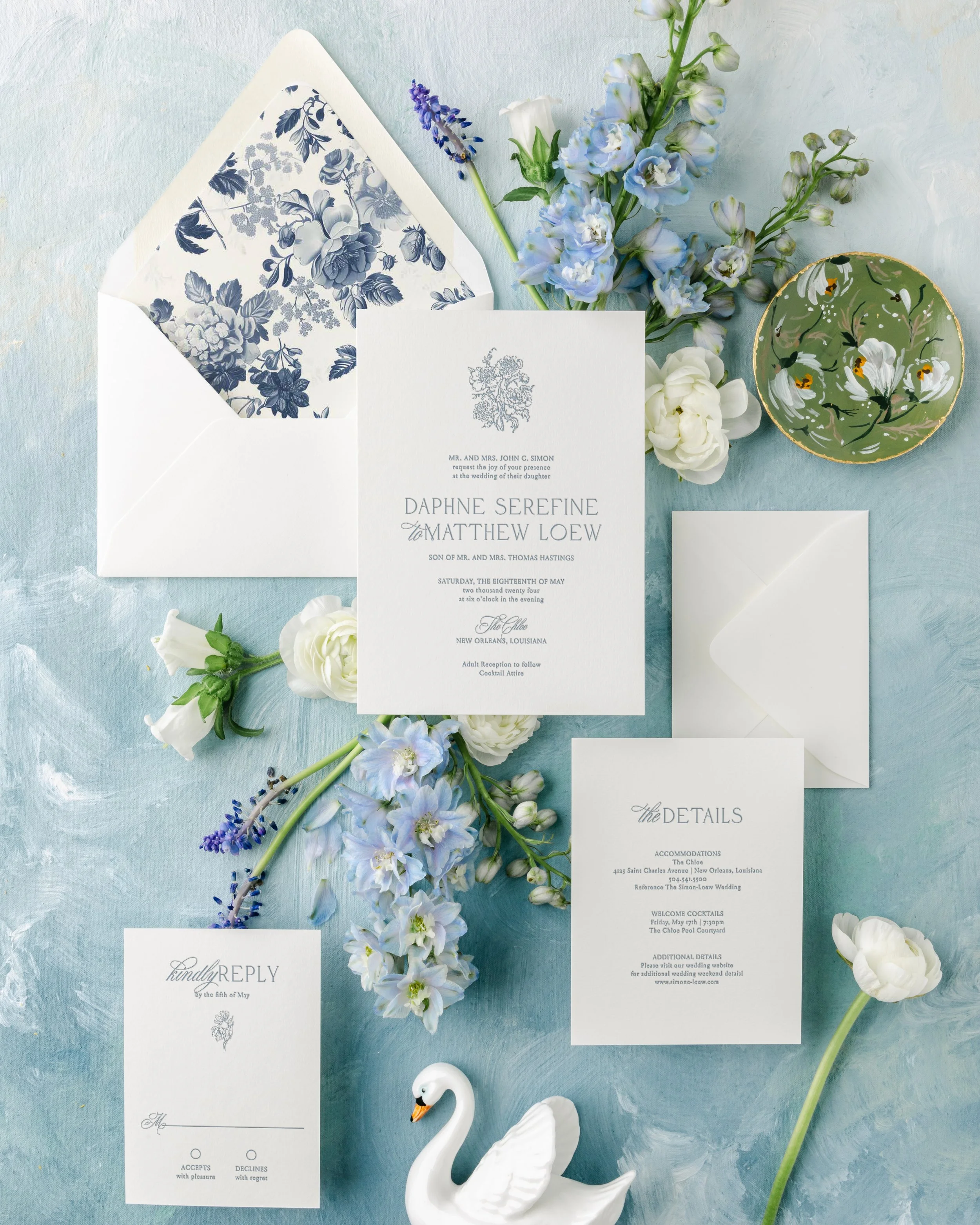 Wedding invitation suite with floral accents, including an envelope with a blue floral liner, a main invitation, a response card, and a details card, surrounded by white and blue flowers, a decorative bowl, and a ceramic swan on a light blue textured surface.