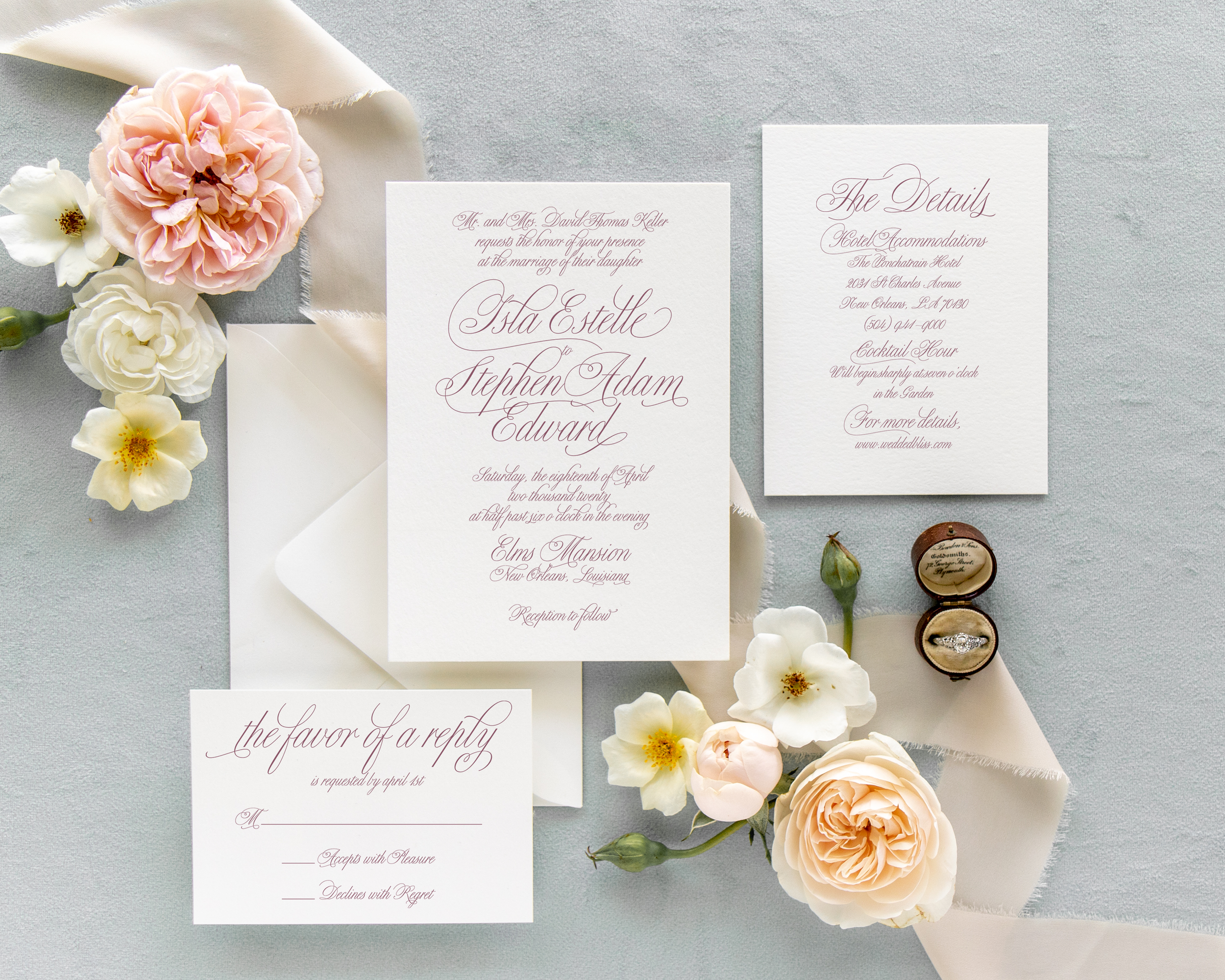Wedding invitation suite with floral accents, featuring pink and white flowers, RSVP card, details card, and a wedding ring in a wooden box on a light background.