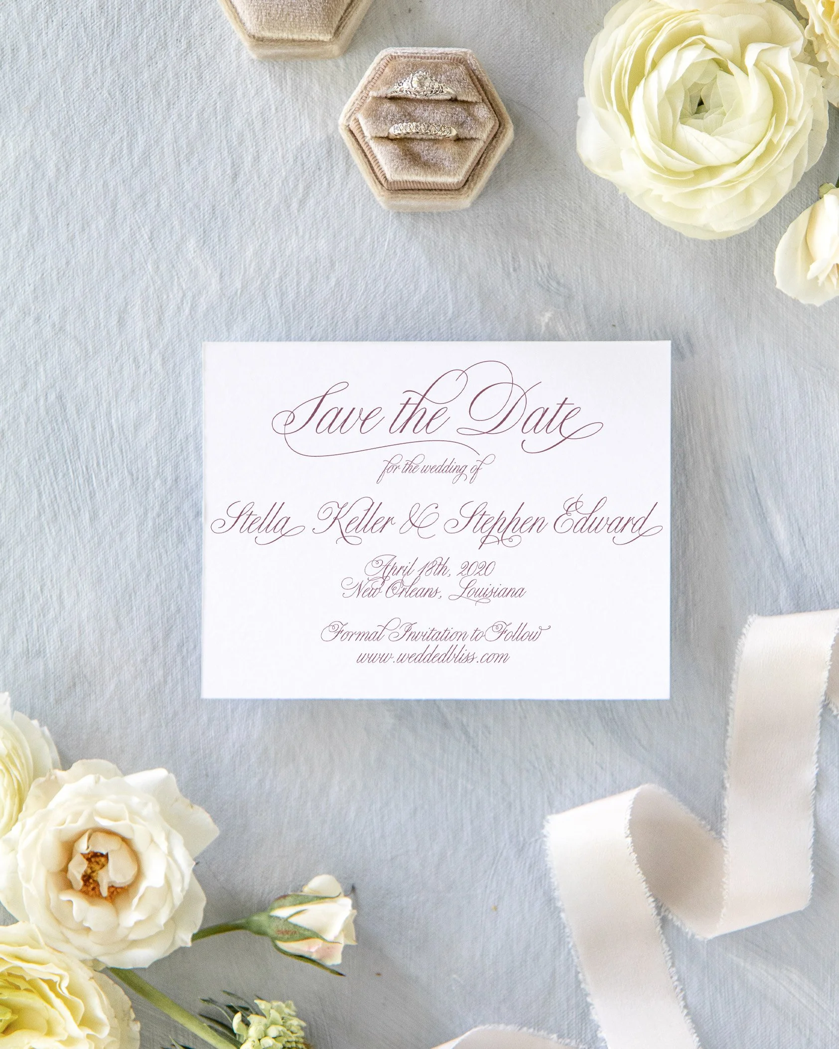A wedding save the date card with elegant cursive writing. Surrounding the card are white flowers, a white ribbon, and a hexagon-shaped jewelry box with rings inside, all arranged on a light gray textured surface.