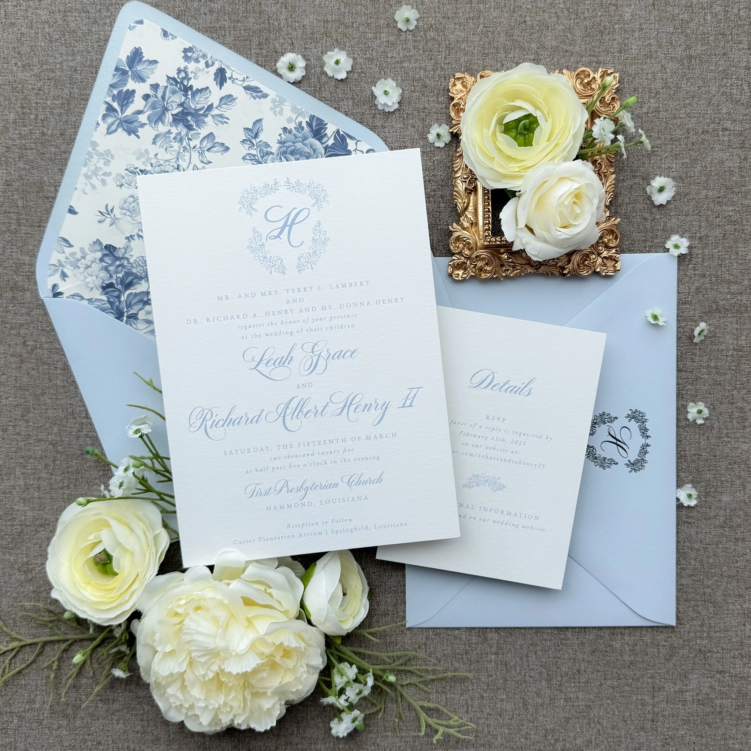 Wedding invitation suite with floral accents, including an Invitation, RSVP card, and details card, accompanied by white roses, green foliage, a decorative gold frame, light blue envelopes, and scattered small white flowers on a textured surface.