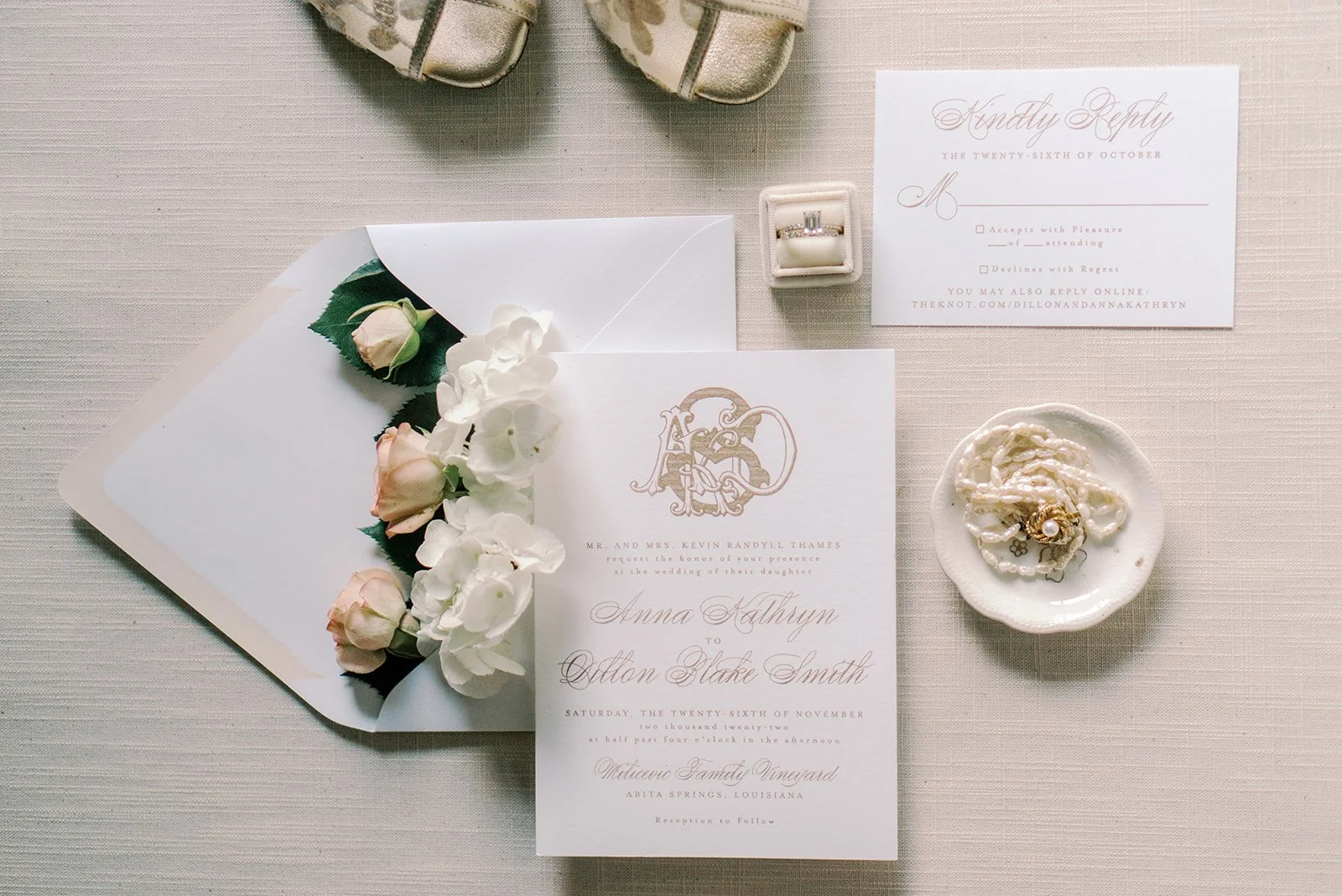 Wedding invitation, floral arrangement, engagement ring, pearl jewelry, and RSVP card on a beige table.