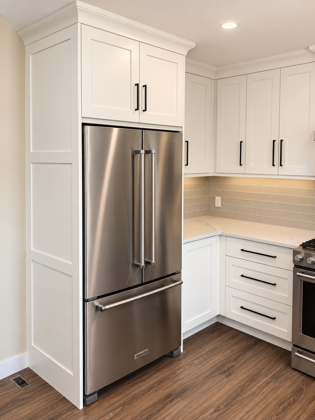 Custom white kitchen cabinetry surrounding a stainless steel refrigerator with stone countertops and warm wood flooring.