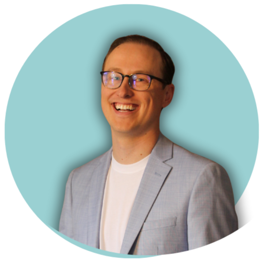 A man smiling, wearing glasses and a light gray blazer over a white shirt, against a light blue background.