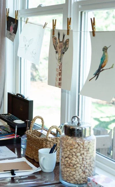 Drawings of a butterfly, a giraffe, and a bird hanging on a clothesline in front of a window inside a room.