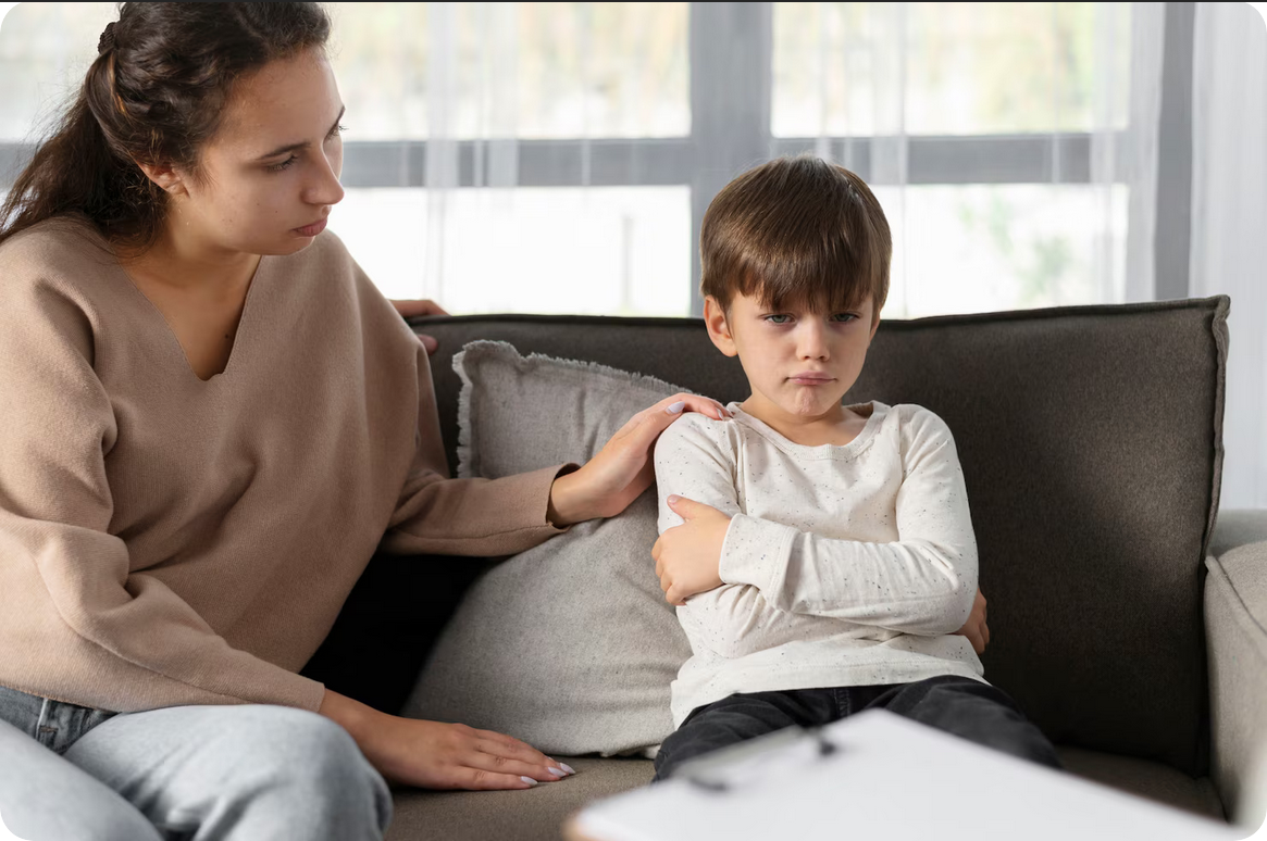 A woman comforts a boy who looks upset, sitting together on a gray couch in a living room.