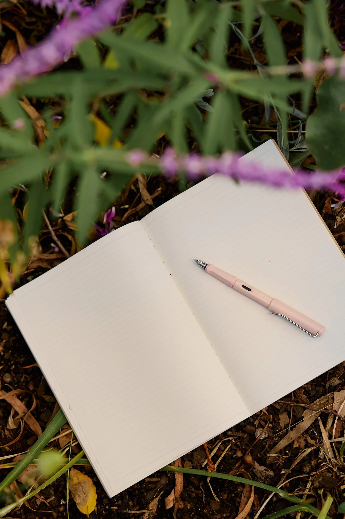 Open blank notebook with lined pages and a pink pen resting on it, placed on the ground among dried leaves, plants, and flowers.