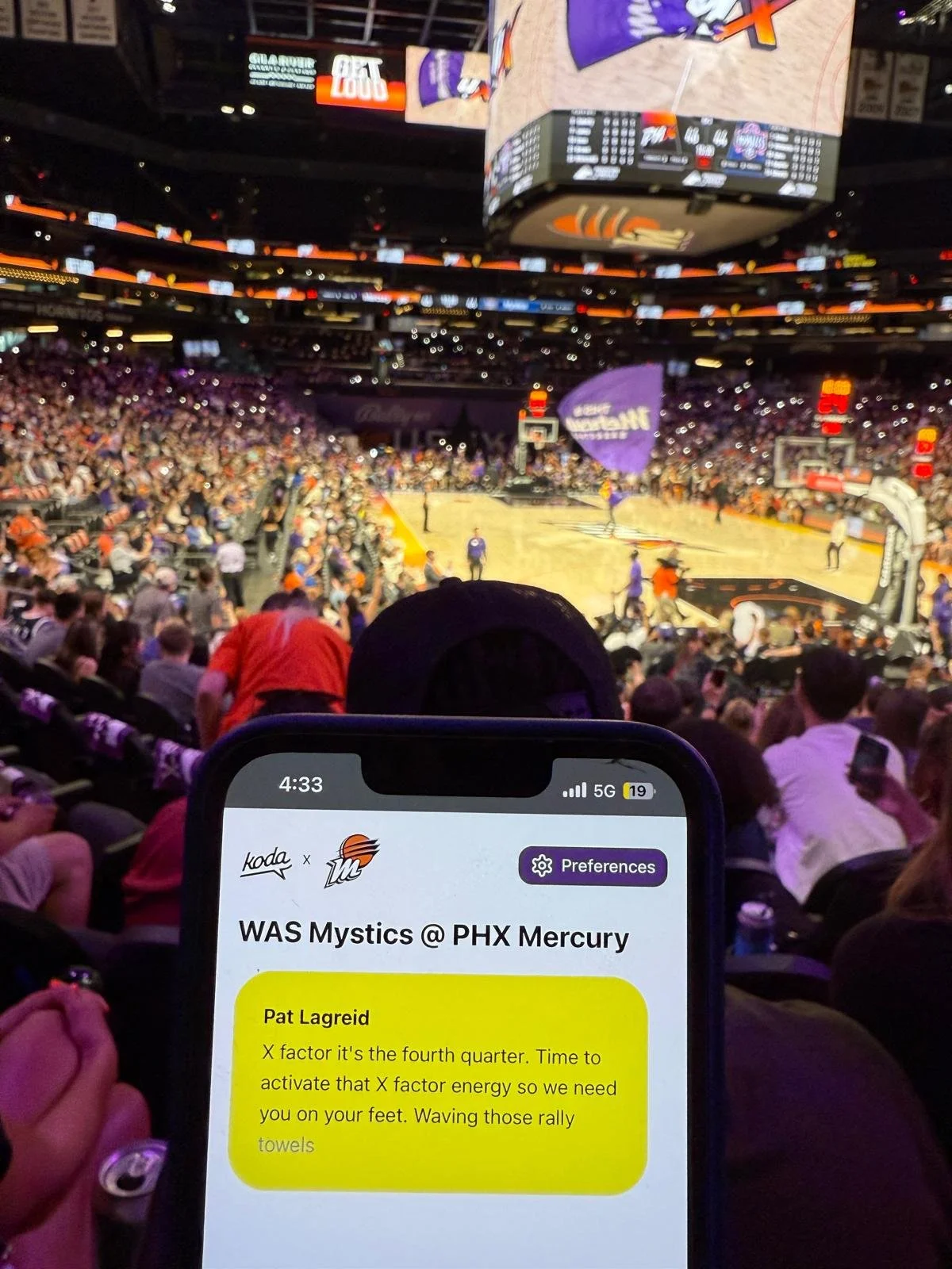 A crowded WNBA game between the Washington Mystics and Phoenix Mercury, with a smartphone in the foreground displaying Koda captions that transcribe the announcer’s words in real time.