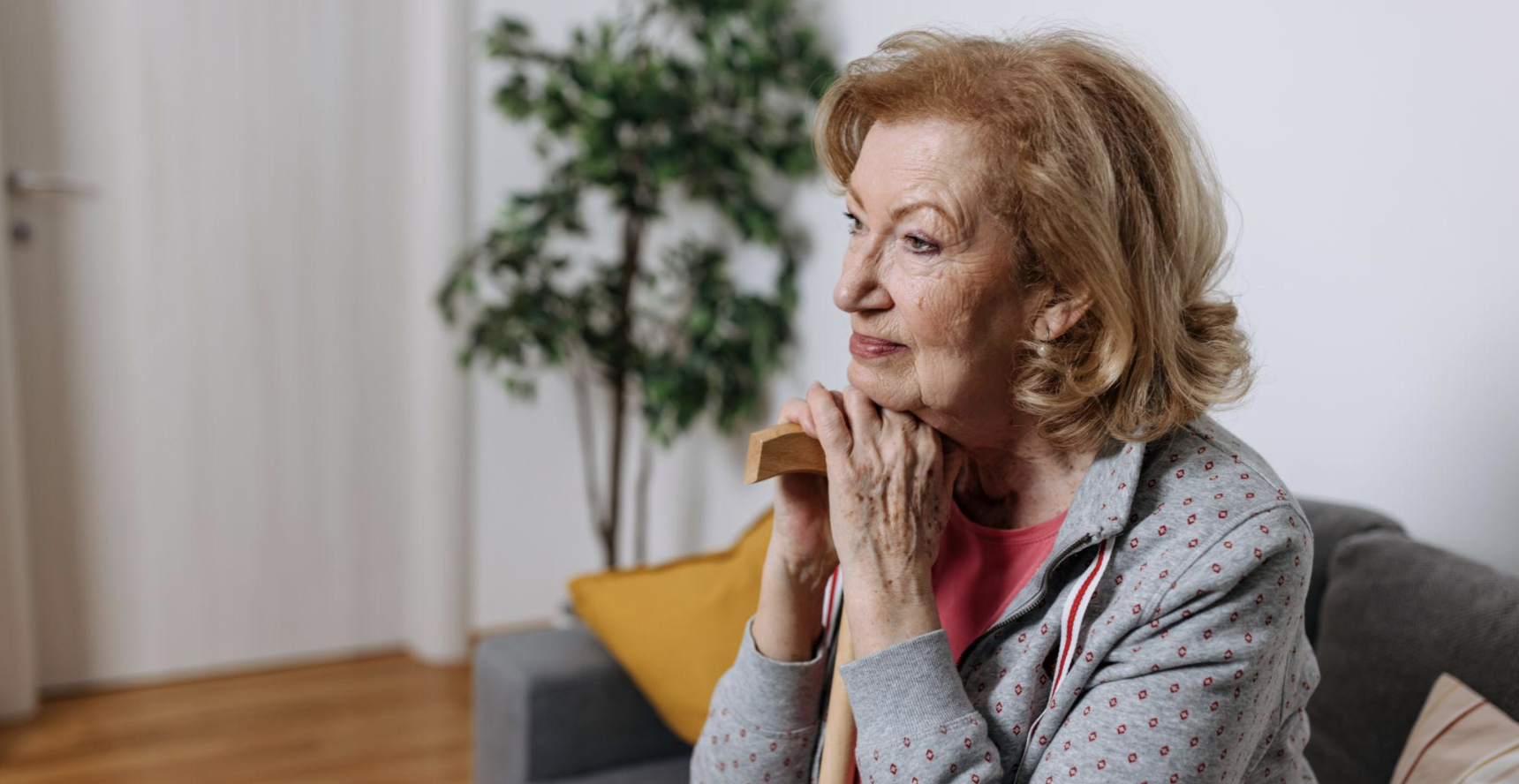 Older woman with red hair sitting on a sofa, resting her chin on a cane, gazing thoughtfully to the side in a cozy living room.