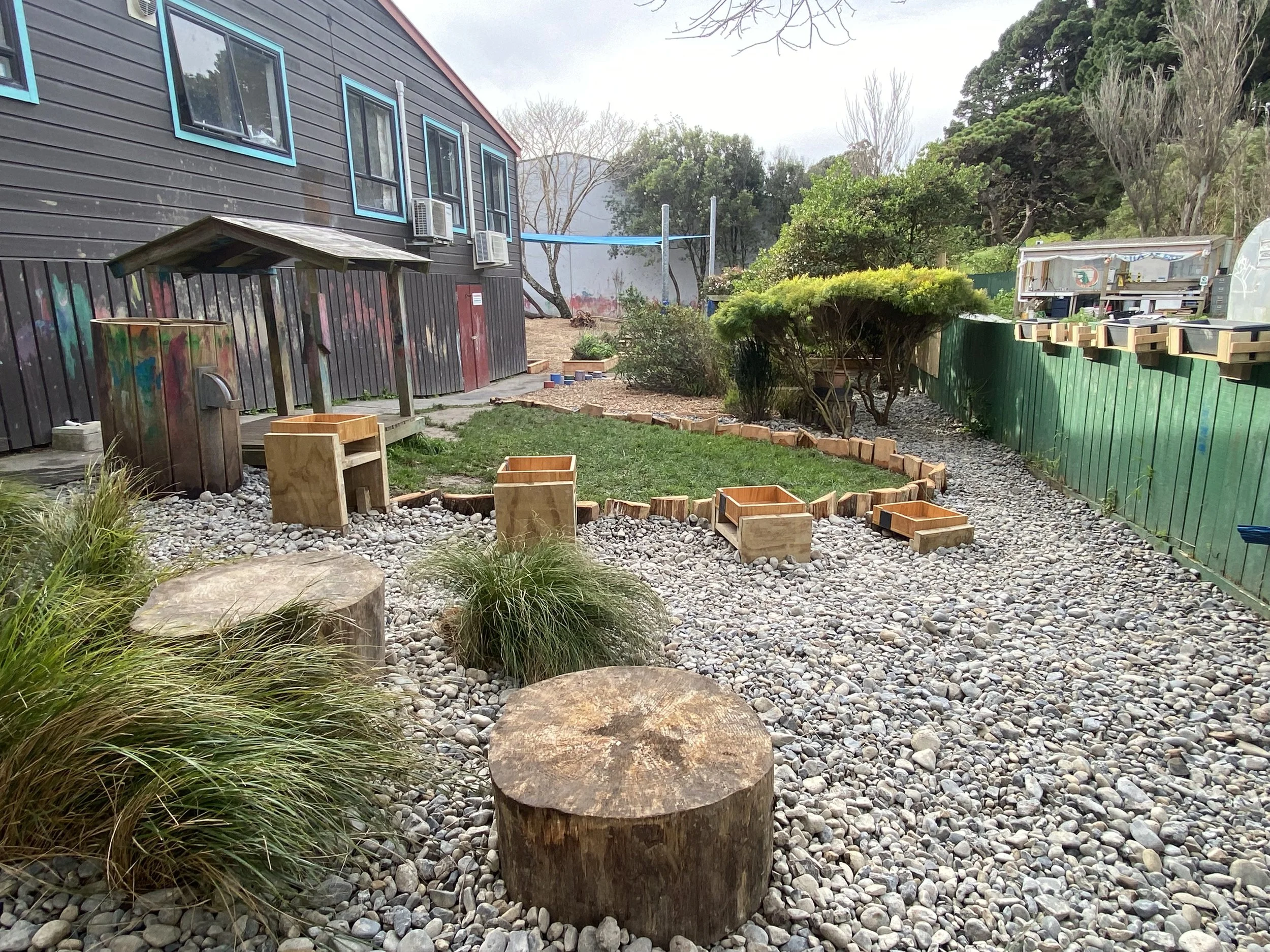 A yard with a gravel ground, large tree stumps, wooden planters, green plants, and a black building with colorful graffiti, along with a green fence on the right side and trees in the background.