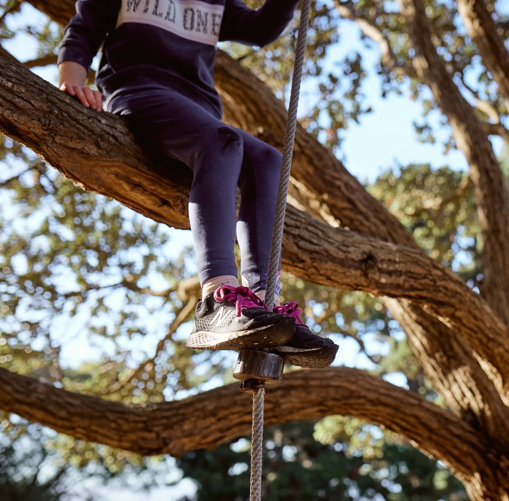 A person wearing black sneakers with pink laces walking on a wooden and rope obstacle in a tree, wearing black athletic clothing.