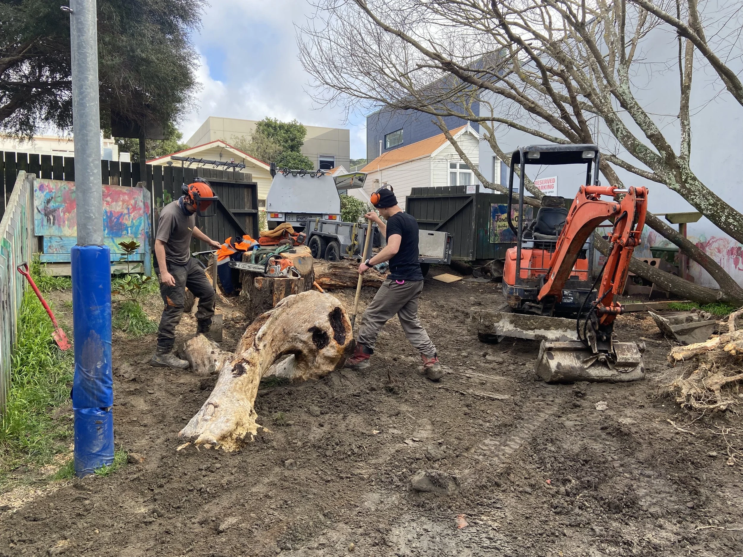 Two workers in hard hats and safety gear are cutting down a large tree on a construction site. The site has a small excavator, a tree stump, and nearby colorful graffiti on the fence. Houses and a truck are visible in the background.