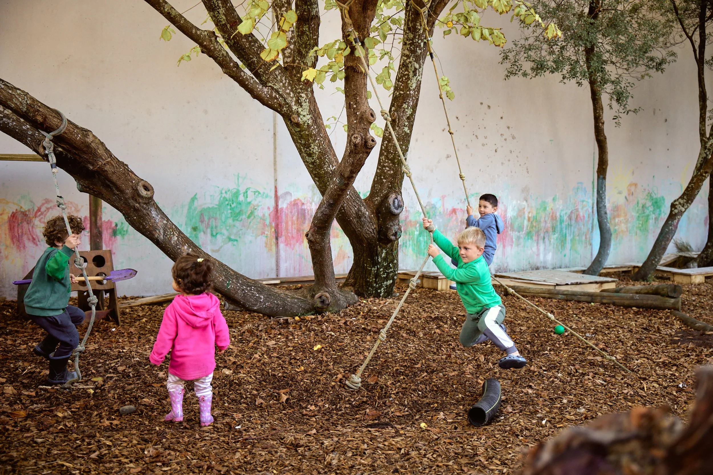 Four children playing on swings and a climbing rope in an outdoor playground with trees and a painted wall in the background.