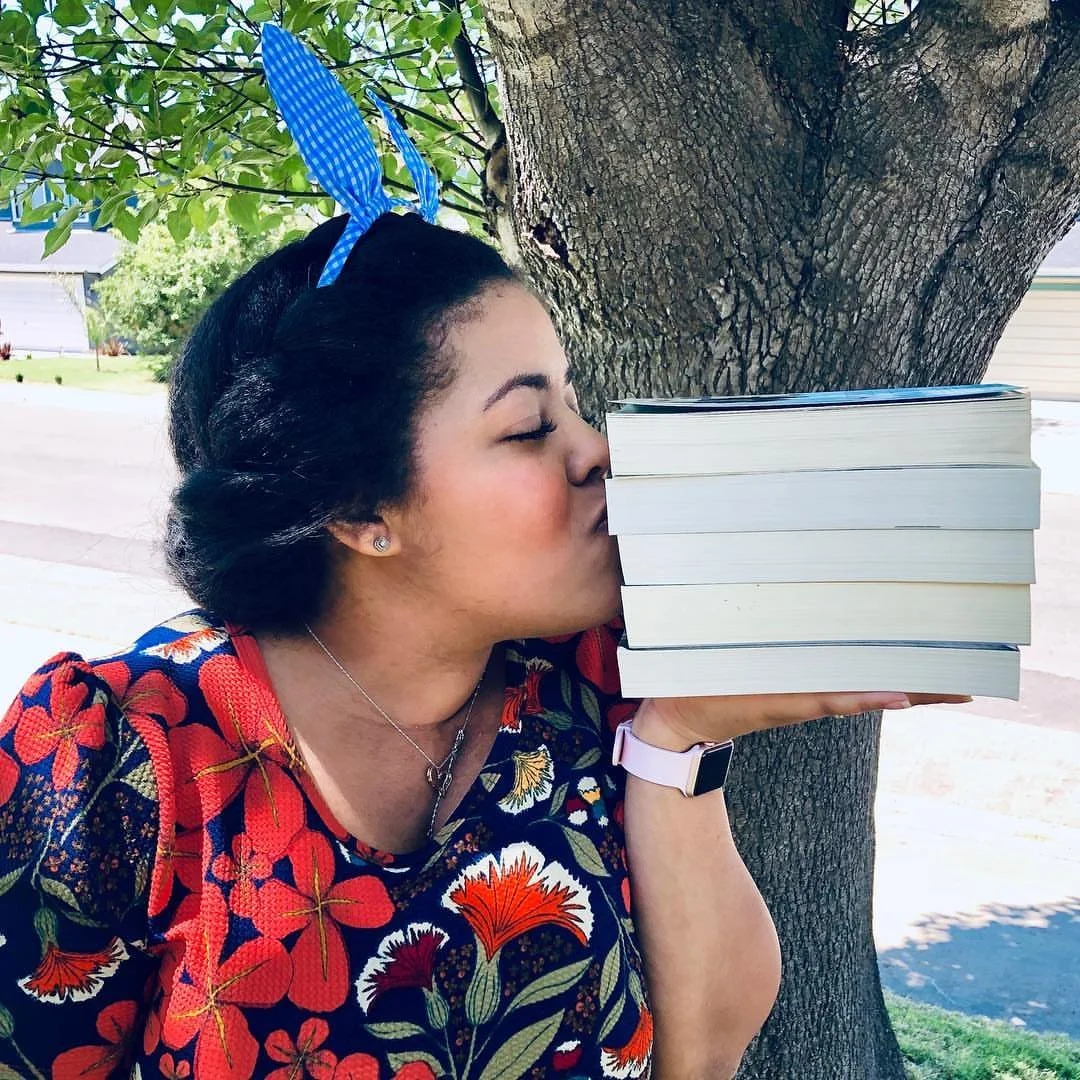 Woman in floral dress kissing book stack in front of a tree.