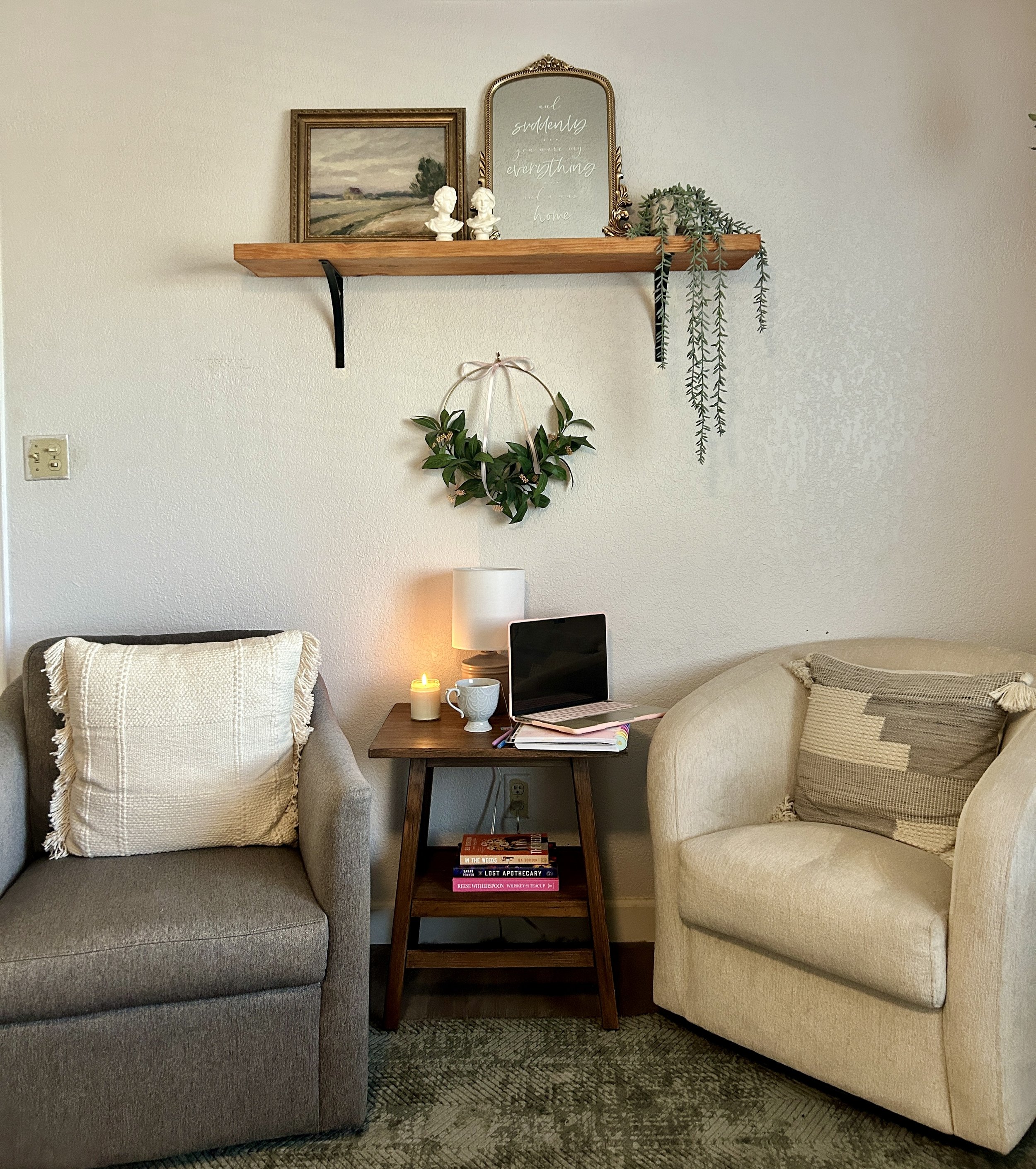Cozy chairs with end table, candle, and laptop work space. Overhead shelf with plant, and welcoming decor. Light academia, books, and tea.
