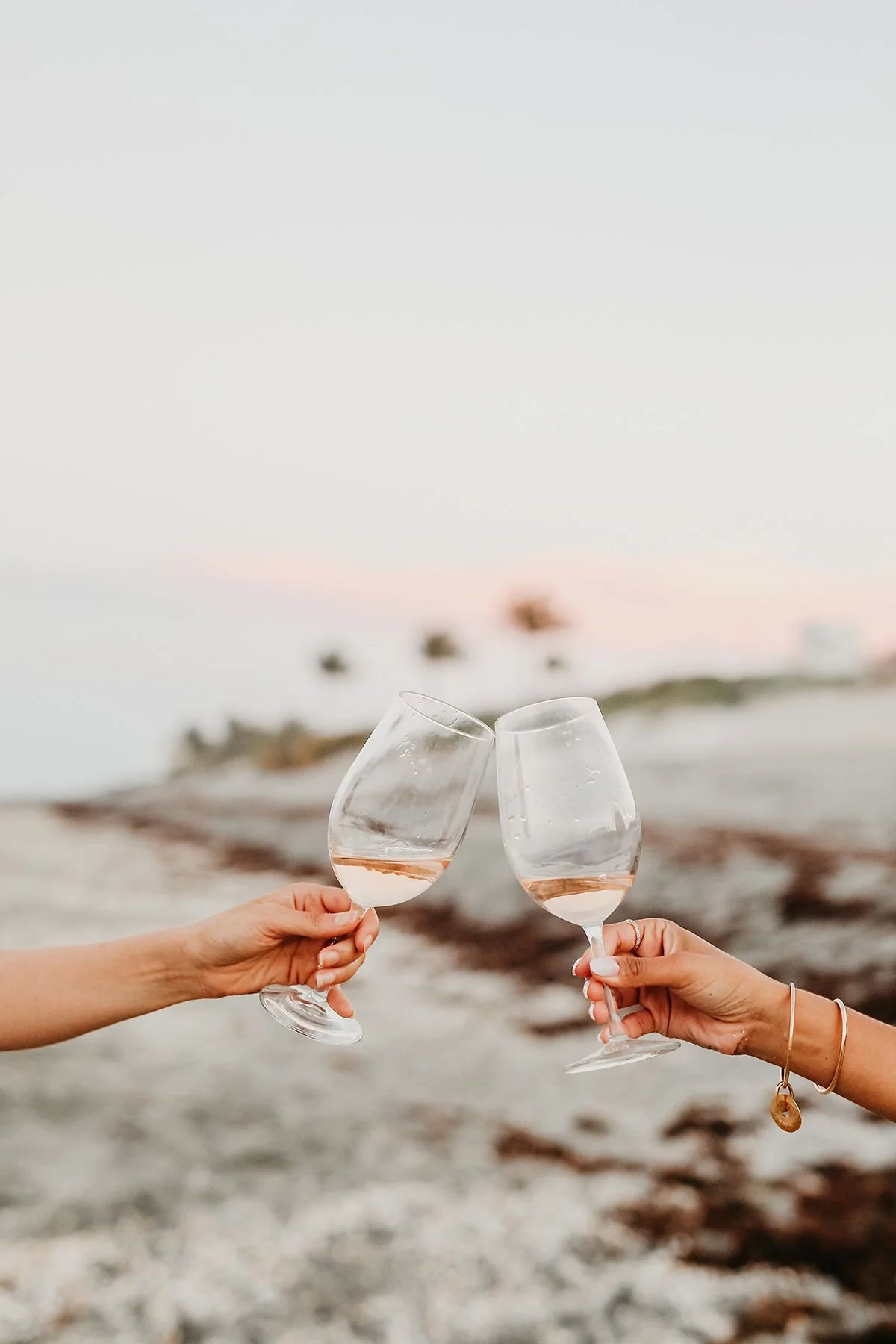 Two women's arms clinking wine glasses in front of a sunset on beach.