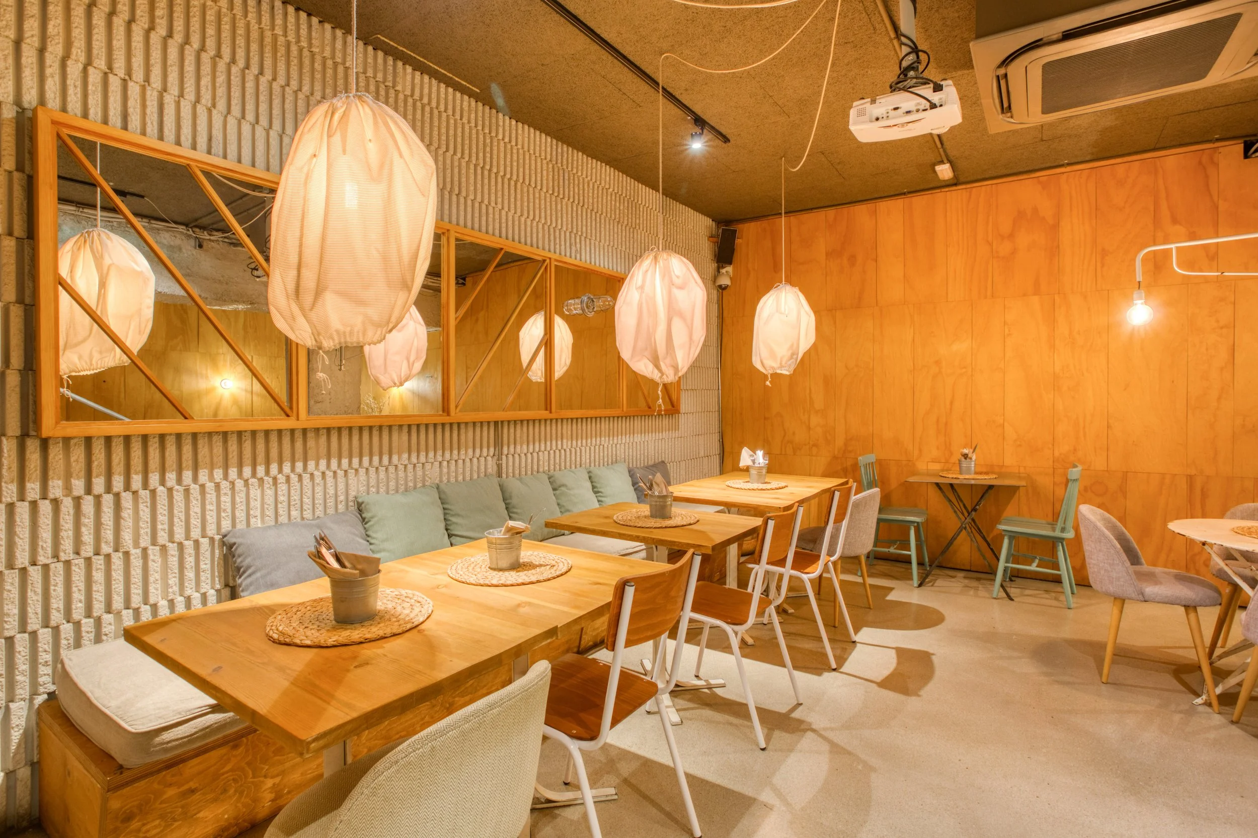 Interior de un restaurante con paredes de ladrillo y madera, lámparas de papel colgantes y mesas de madera con sillas variadas, decoradas con centros de mesa de jarrones con utensilios de cocina.