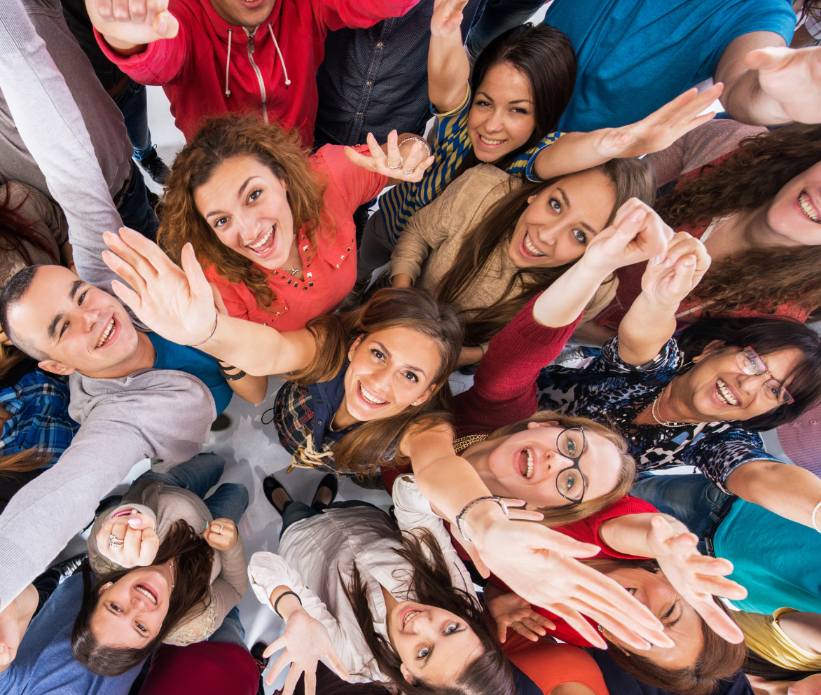 Photo taken above of many people smiling and looking up at camera with their arms reaching up over their heads.