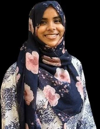 A young woman wearing a navy blue floral hijab and matching dress, smiling against a black background.