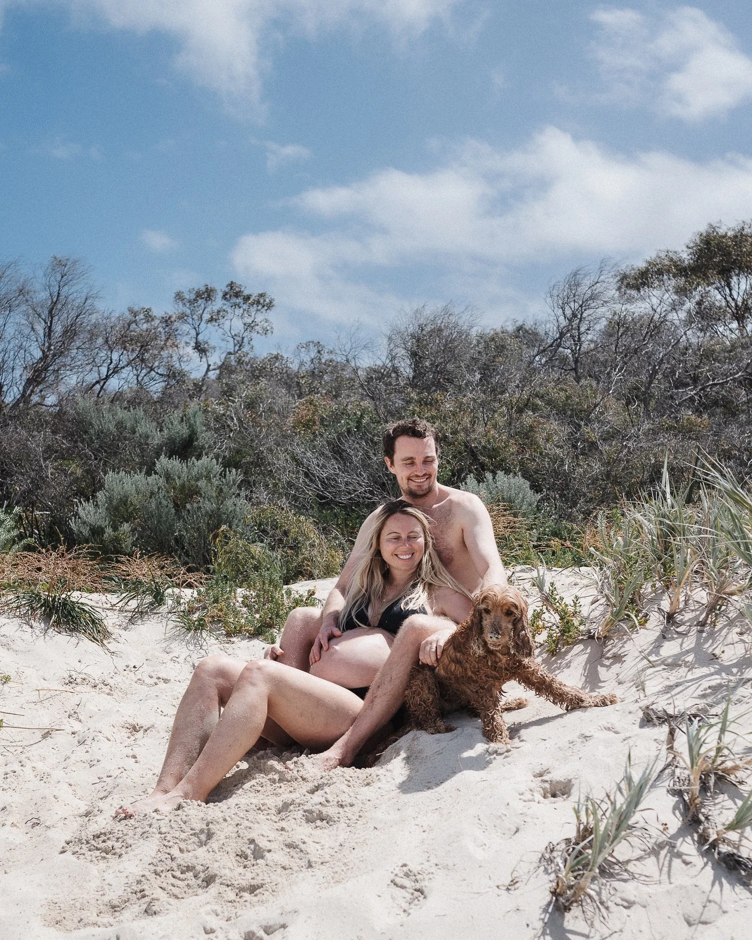 A couple and their dog sitting on sandy beach with bushes and trees in the background under a partly cloudy sky.