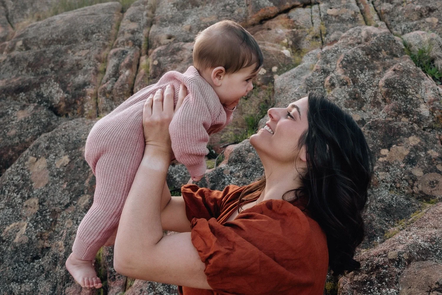 A woman with dark hair smiling and holding a young child in a pink outfit, with a rocky background.
