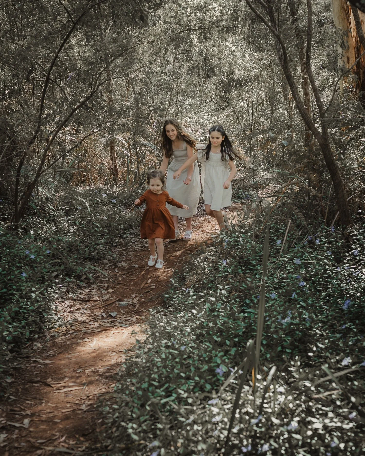 Three young girls, one toddler, walking on a forest trail surrounded by dense greenery and trees.