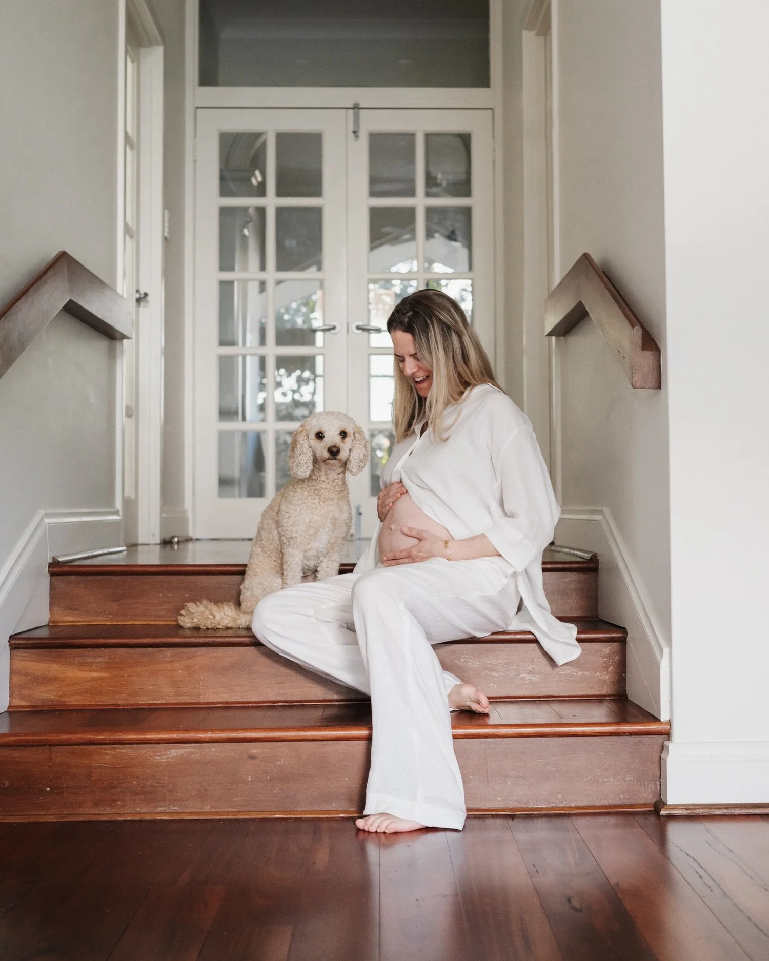 Pregnant woman sitting on wooden stairs with a dog beside her, smiling and looking down at her belly.