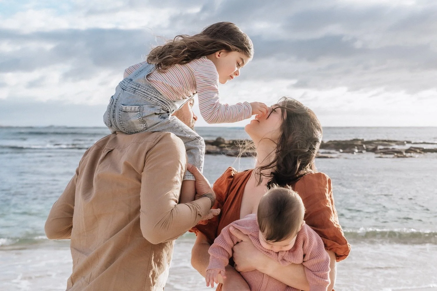 A family of four enjoying time at the beach. The father is holding a toddler, the mother is holding a baby, and a young girl is touching the mother's nose and leaning on her.