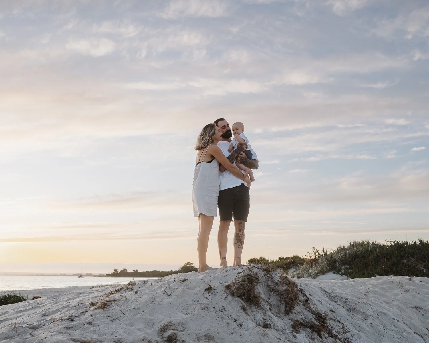 A family of three, consisting of a man, woman, and baby, standing on a sandy beach at sunset, embracing each other.