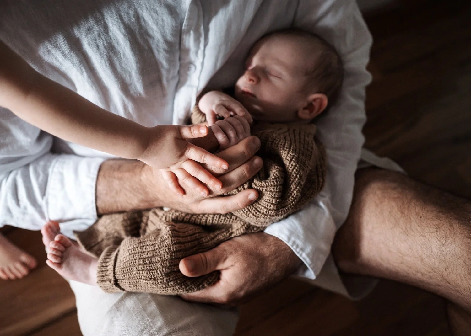 A baby lying on an adult's lap, holding hands with a child's hand, wrapped in a brown knitted blanket.