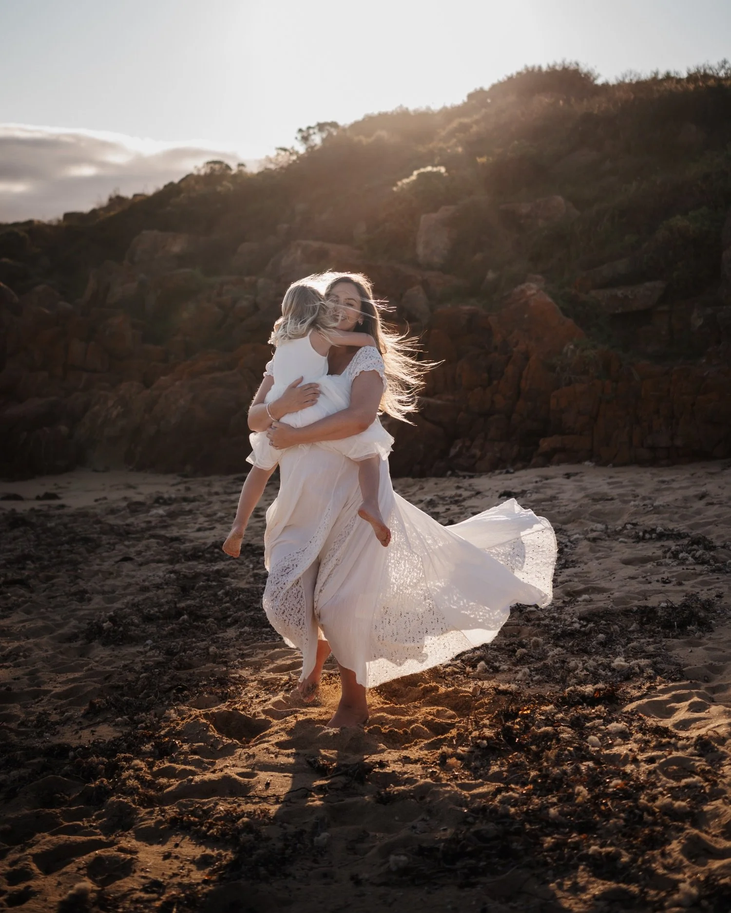 A woman in a white flowing dress running barefoot on the beach while holding a young girl in a white dress who is hugging her.