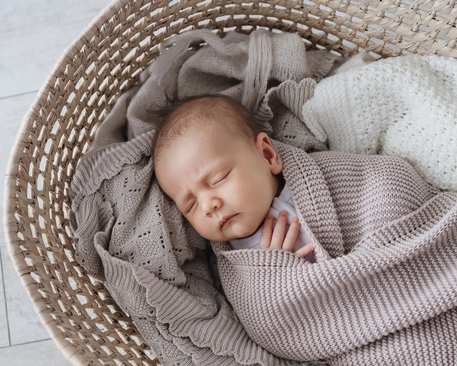 A sleeping baby in a wicker bassinet, wrapped in a beige knitted blanket with other soft blankets around, resting peacefully.