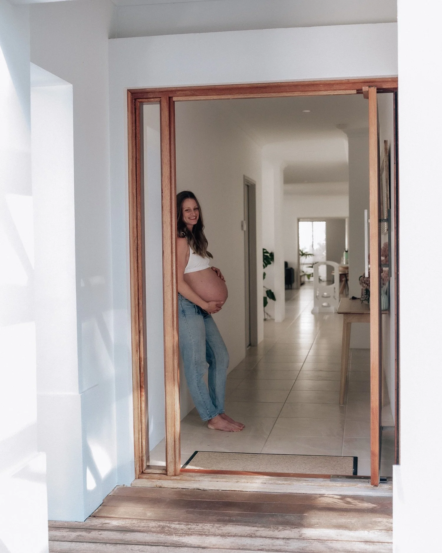 Woman with a pregnant belly standing barefoot in a doorway, smiling, inside a bright, modern home with white walls and wooden accents.