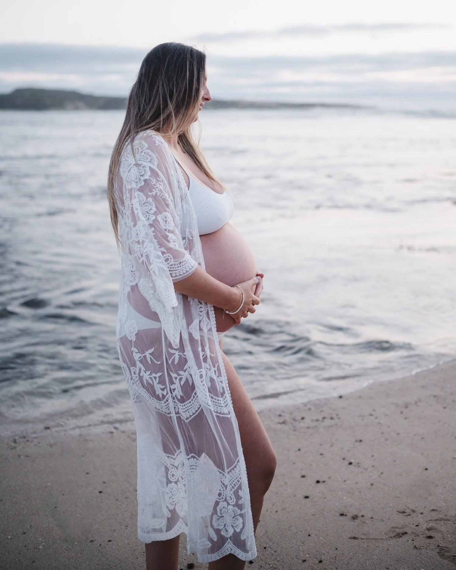 Pregnant woman in white lingerie with a lace cover-up standing on the beach holding her belly with one hand, looking down, with the ocean in the background.