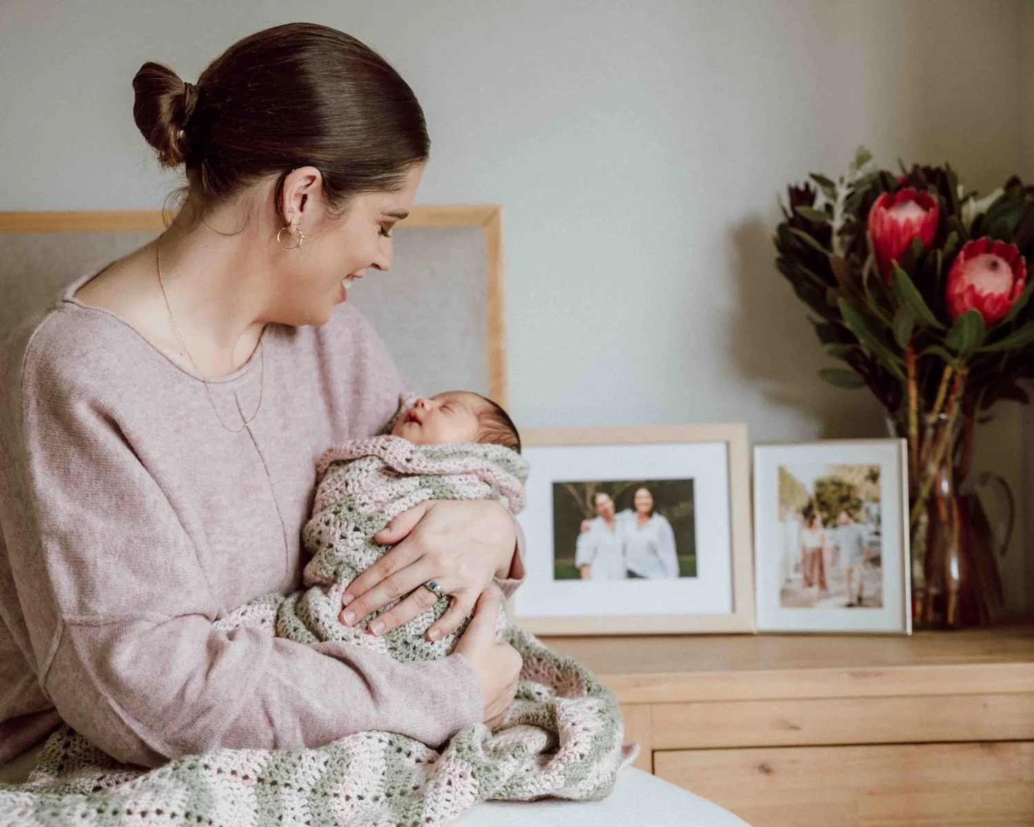 A woman holding a sleeping baby wrapped in a knitted blanket, smiling and looking at each other in a cozy room with framed family photos and large flowers on a wooden surface in the background.