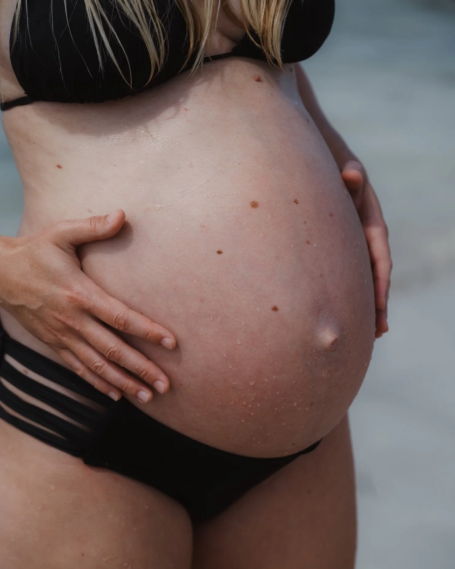 Close-up of a pregnant woman's swollen belly with water droplets, moles, and a belly button, as she touches her stomach with her left hand at the beach.