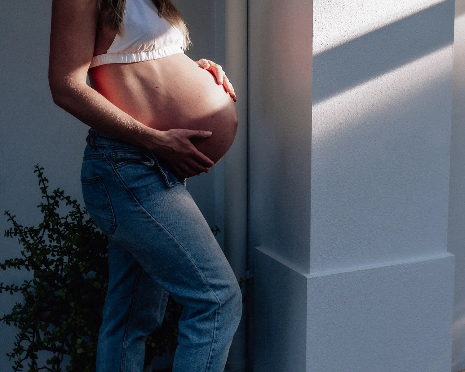 A pregnant woman standing outdoors, touching her belly, with sunlight and shadows cast on a white wall.