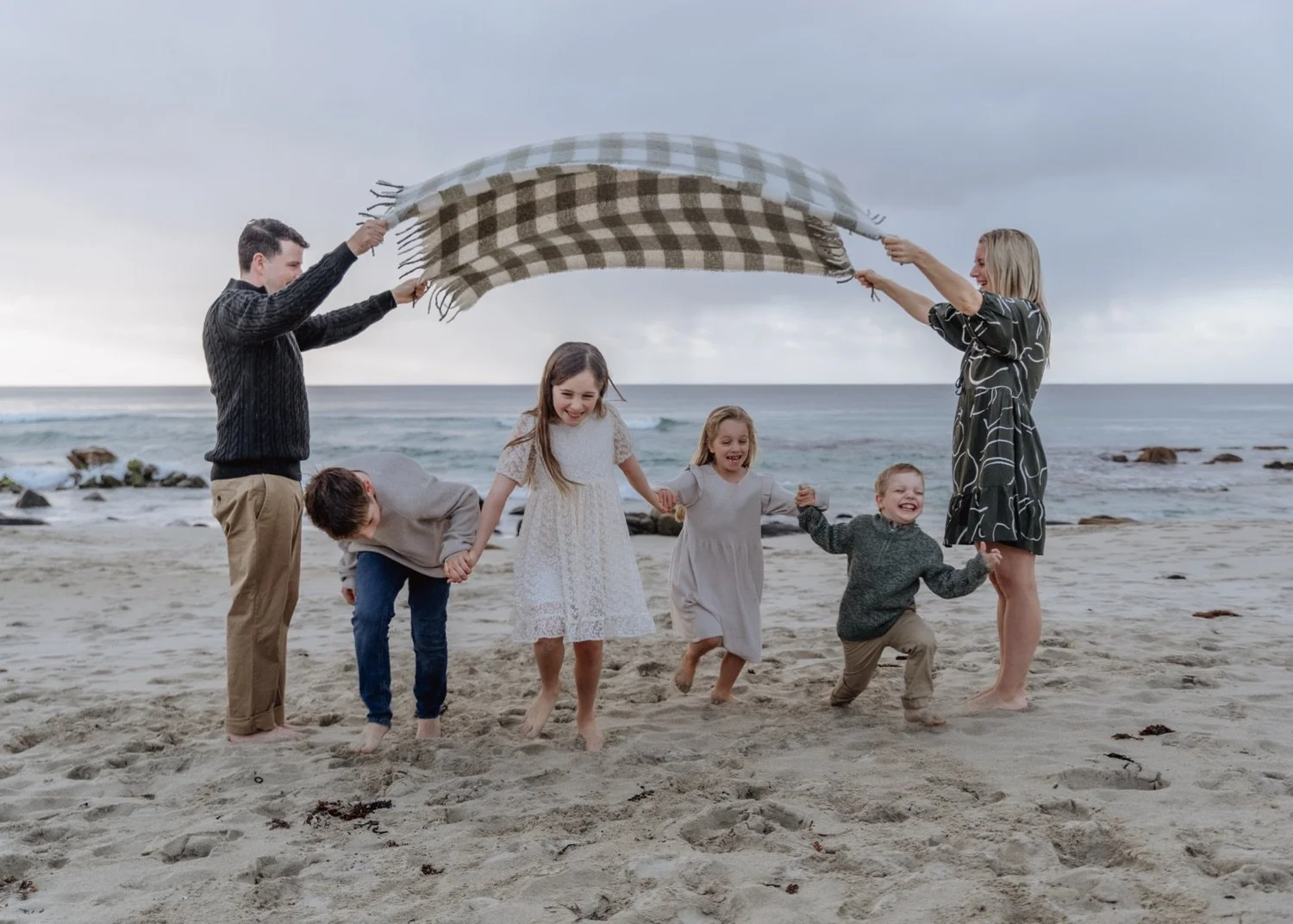 Family of six playing on the beach with a blanket over their heads, holding hands and smiling, with the ocean and cloudy sky in the background.