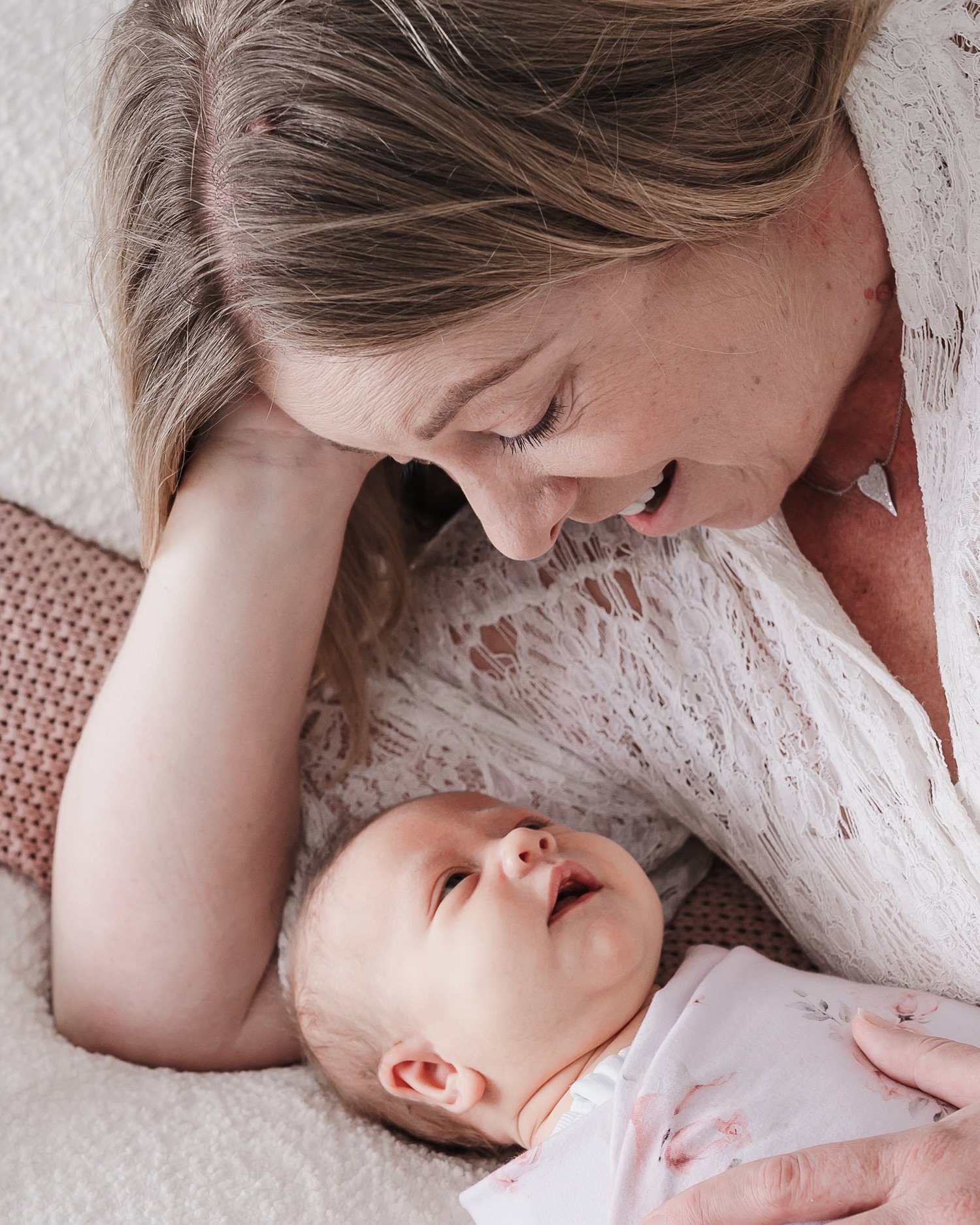 A woman with blonde hair and a white lace top lies on her side, looking down and smiling at a baby girl with light skin and dark hair, who is lying on her back and smiling.