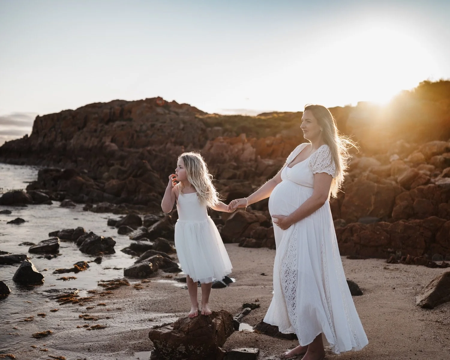 A pregnant woman and a young girl holding hands by the beach at sunset, both dressed in white dresses.