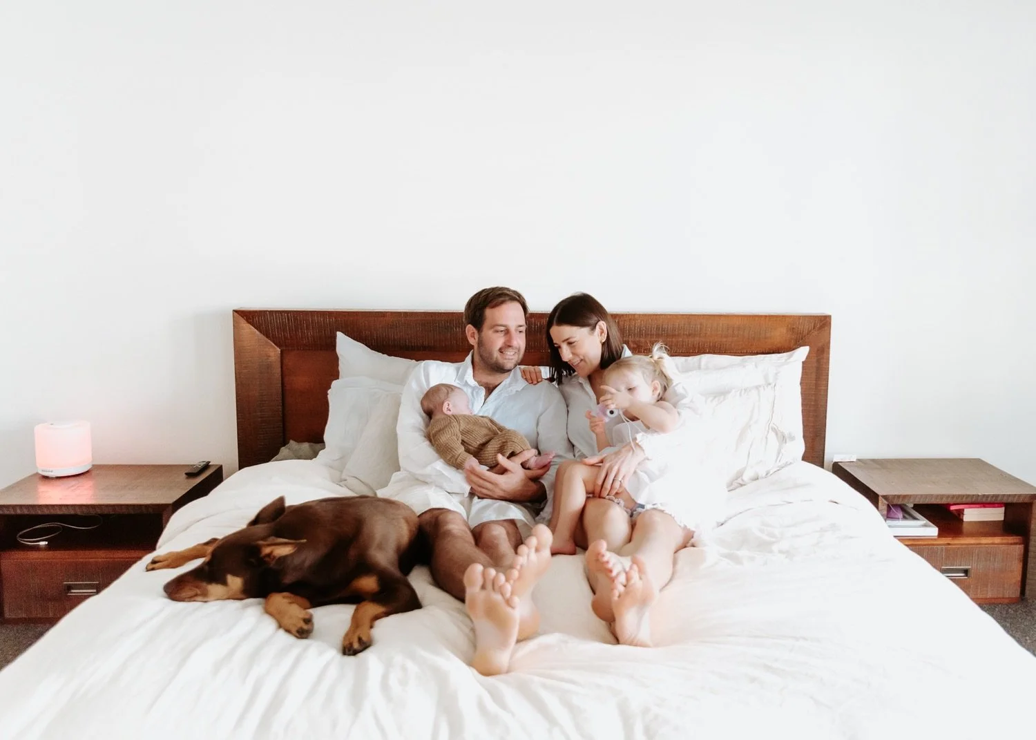 A family of four, including a father, mother, and two young children, sitting on a bed with a brown dog resting nearby. The father is holding a baby, and the mother is playing with a toddler. The scene is bright and cozy with minimal decor.