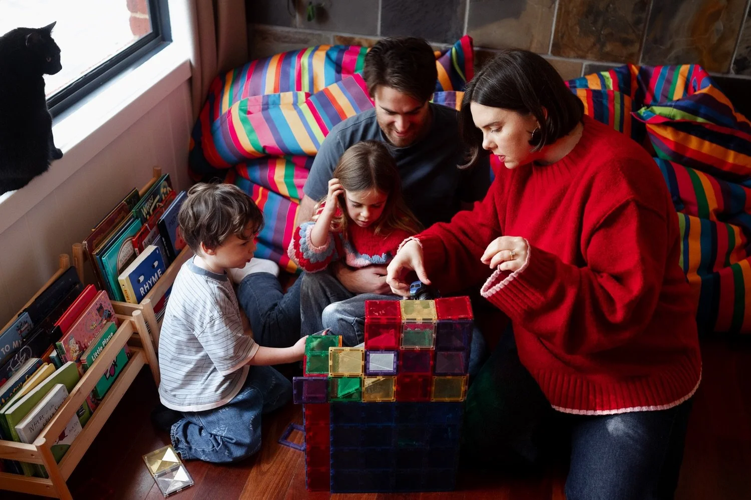 Family of four, including a mother, father, and two children, playing with colorful magnetic building blocks in a cozy living room with a rainbow-striped couch and a bookshelf near a window.